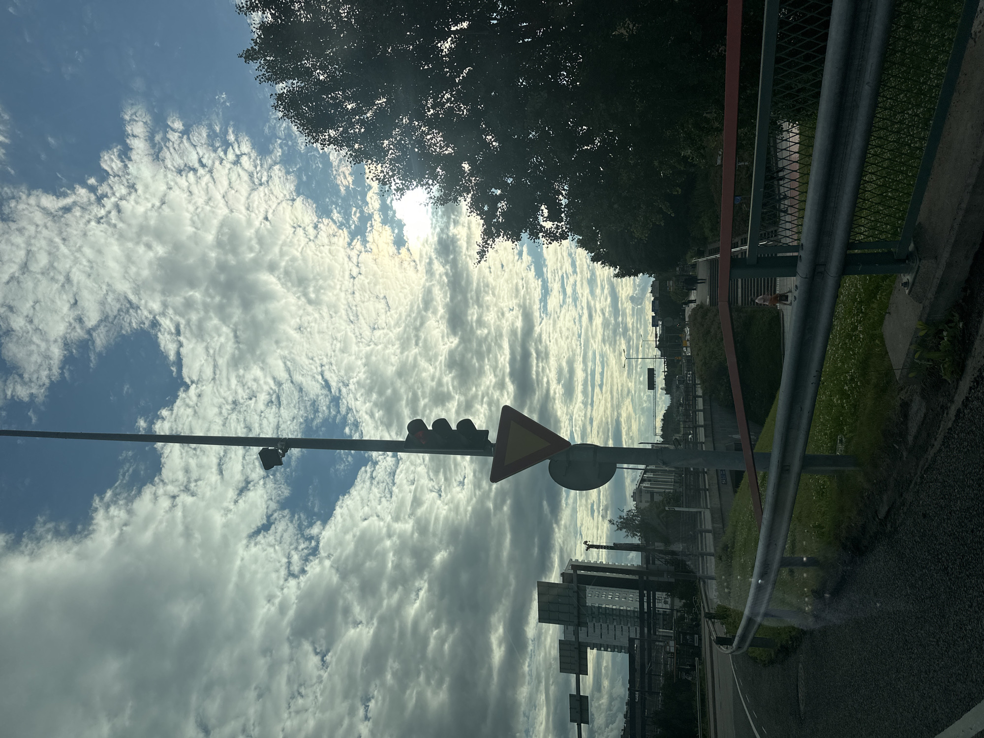 This media file depicts an outdoor scene, likely on a street or roadside, oriented vertically. The sky is filled with dramatic, textured clouds, with patches of blue sky peeking through. Sunlight filters through the clouds, suggesting it is daytime, possibly late morning or afternoon. The sun's glare is visible in the upper portion of the image.
In the foreground, a traffic light with three red lights is prominently displayed on a pole. Next to the traffic light is a yellow triangular warning sign with a red border, indicating a hazard or caution ahead. A circular sign is also visible beneath the triangular sign. A metal guardrail with a red horizontal bar runs along the right side of the frame, separating the roadway from a grassy area.
Further in the background, trees with dense foliage form a dark canopy. Buildings, including what appear to be multi-story structures, are visible in the distance, suggesting an urban or suburban environment. A crane is also visible in the background, hinting at ongoing construction. The ground is paved, with a white line marking a road. There are some blurry reflections on the surface in the lower right part of the image, possibly from the perspective of being inside a vehicle. The overall impression is a bright but slightly overcast day.