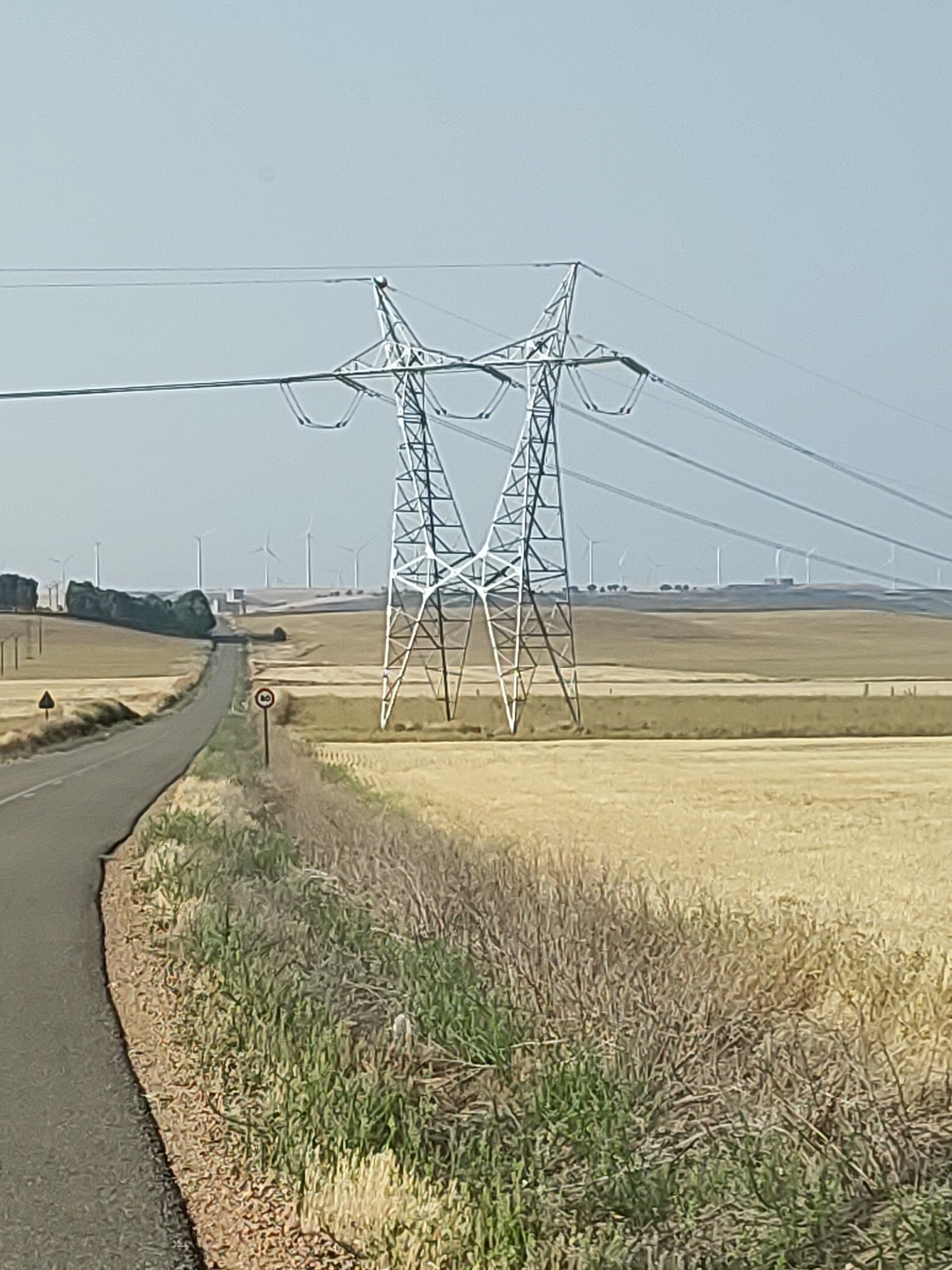 The image depicts a rural landscape in the vicinity of Torremormojón, Spain. A prominent electrical pylon stands in the foreground, supporting high-voltage power lines that stretch across the frame. To the left, a paved road curves through a field of dry, golden crops, likely wheat or barley, ready for harvest. In the distance, a row of wind turbines dots the horizon, indicating a renewable energy farm. The sky is a pale, hazy blue, characteristic of a warm, sunny day. A circular speed limit sign is visible on the side of the road. The overall scene conveys a sense of open space and agricultural activity under a clear sky.