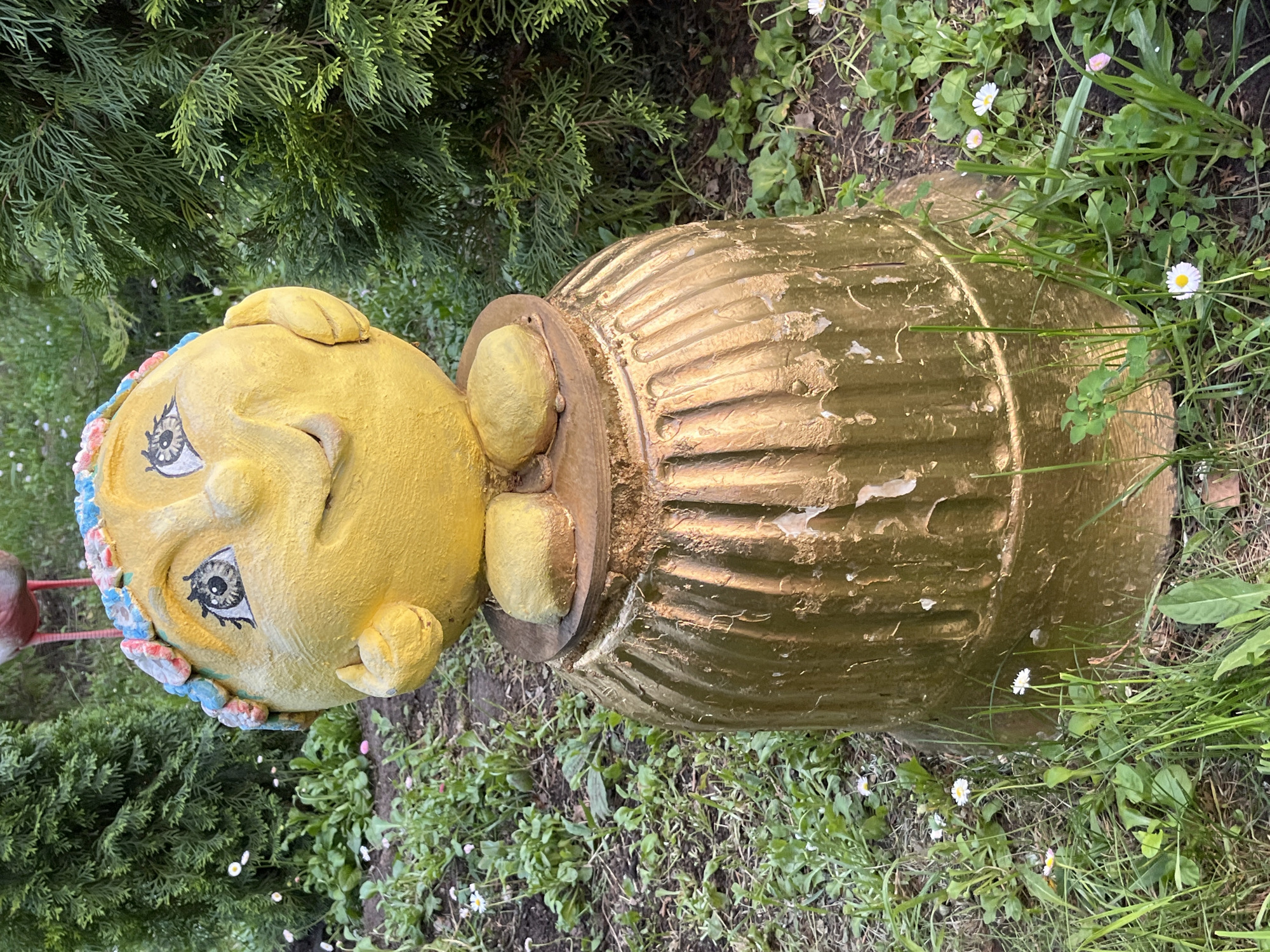 A garden decoration in Chaikovsky, Russia, consisting of a golden urn with a yellow head on top is partially visible. The head is adorned with a colorful floral crown. The decoration sits amidst grass, clover, and small white flowers next to a green bush.