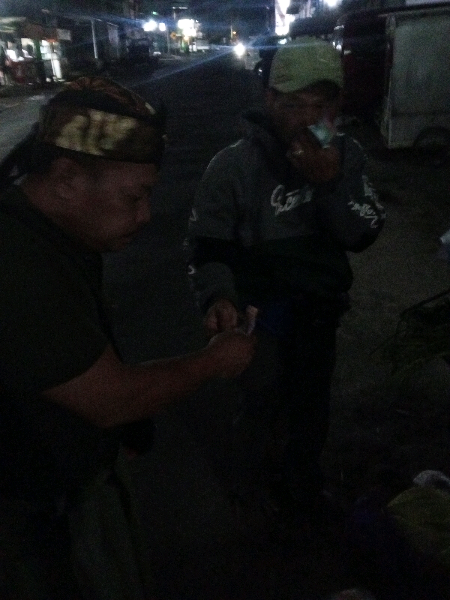The image depicts two individuals outdoors at night, possibly on a street in Gentan, Indonesia. The person on the left wears a dark shirt and a patterned head covering. Their face is partially visible. The person on the right, wearing a light green cap and a dark hooded sweatshirt, is holding a cigarette to their mouth and appears to be smoking. Their face is also visible. The background shows a dimly lit street scene with buildings and vehicles, suggesting an urban environment. The lighting is low, creating shadows and a grainy effect.