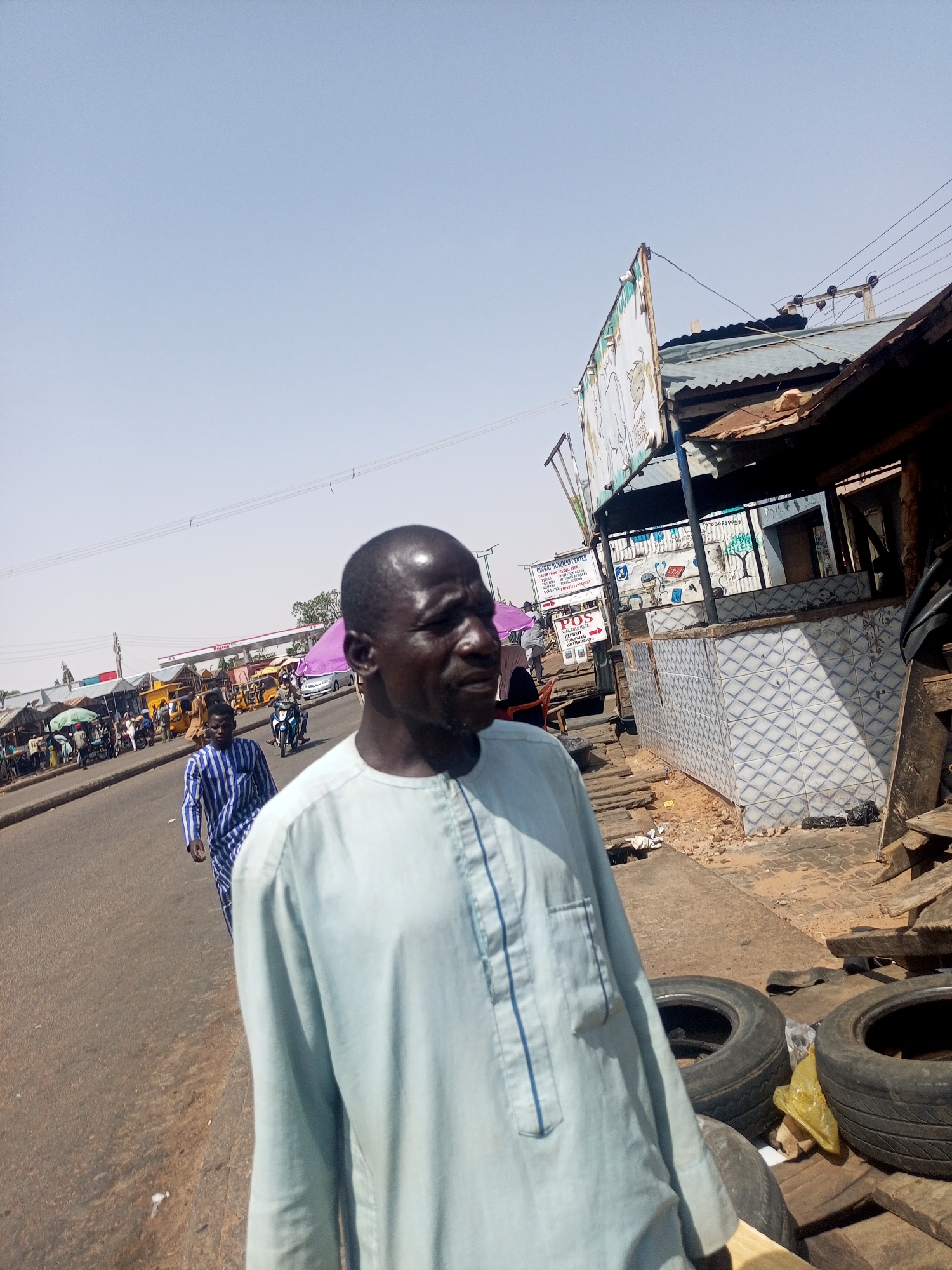The image depicts a street scene in Gombe, Nigeria. An adult male in a light blue tunic stands prominently in the foreground, looking to his right. Another man in a blue and white striped robe walks down the street behind him. In the background, there is a gas station, motorcycles, and people under umbrellas. A storefront with a faded sign is to the right. Three tires are on the sidewalk by the storefront.