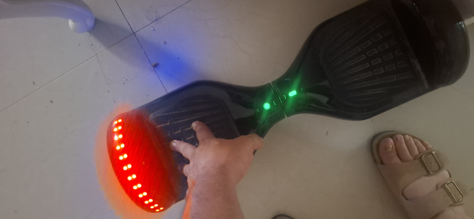 A person's hand is seen resting on the side of a black hoverboard. The hoverboard has several green LED lights illuminated in the center and a strip of red LED lights on the side closest to the viewer. Blue LED lights are also visible on the floor near the hoverboard. A person's foot wearing a light-colored sandal is visible to the right of the hoverboard. The hoverboard is positioned on a light-colored tiled floor, possibly in an indoor setting.