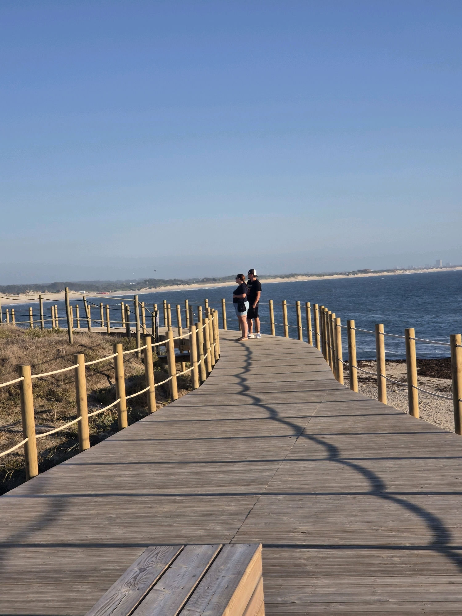 The image depicts a wooden boardwalk leading towards the ocean in Esposende, Portugal. Two individuals, a man and a woman, stand on the boardwalk facing the water, with the man appearing to take a photo of the woman. The boardwalk is bordered by a sandy dune area with vegetation and a wooden post-and-rope fence. The ocean is calm, with a distant shoreline visible. The sky is clear and blue. The scene is set during daylight hours.
