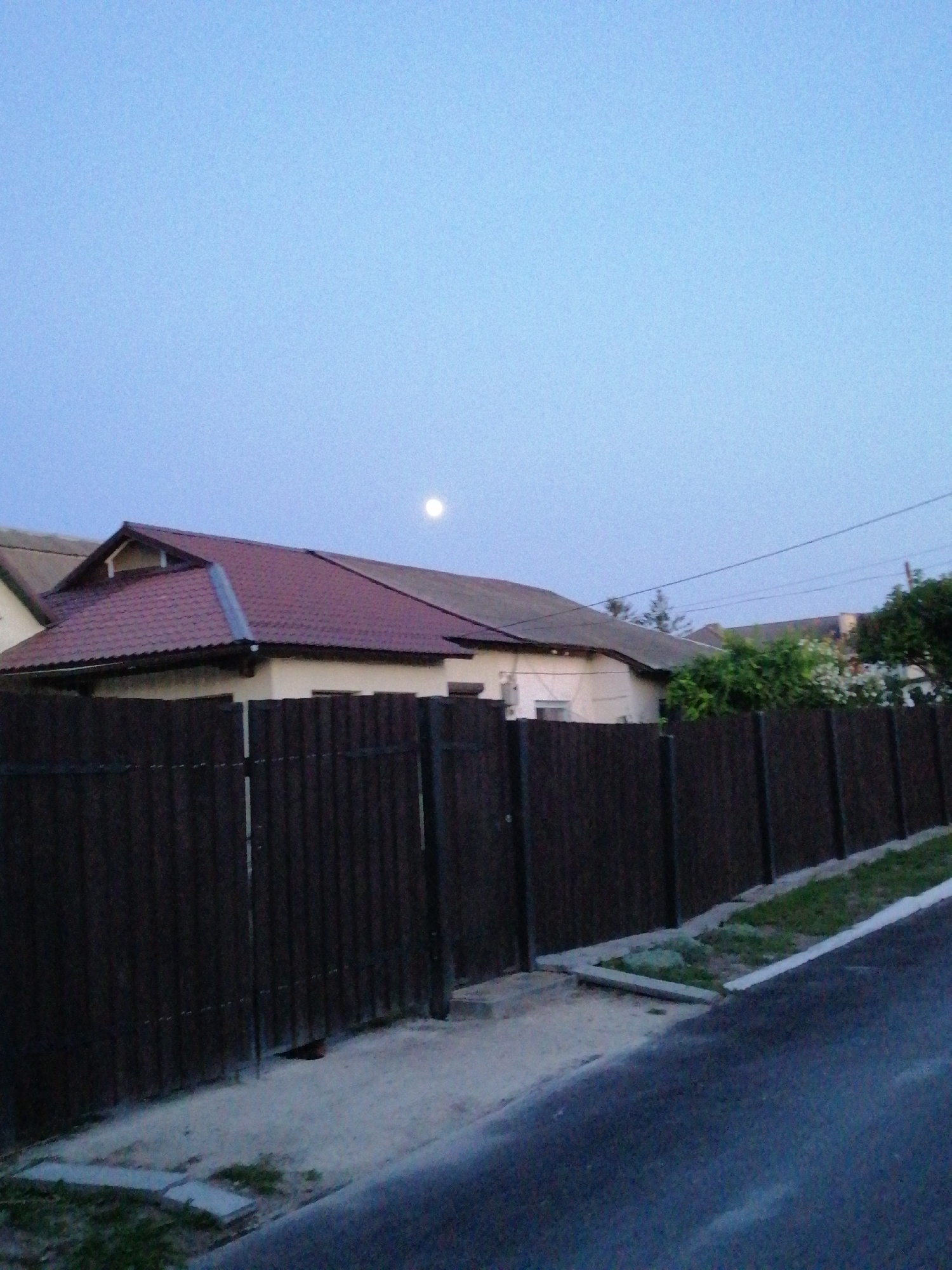 The image depicts a scene at dusk in Lanivtsi, Ukraine, with a pale blue sky dominating the upper portion. A round, faint moon is visible in the sky above a series of residential buildings. The buildings have dark-colored, corrugated metal roofs, and cream-colored walls. A dark brown, vertical metal fence runs along the foreground, separating the street from the properties. The street itself appears to be paved with dark asphalt, and a narrow strip of grass and concrete edging is visible between the street and the fence. The overall atmosphere is calm and suburban.