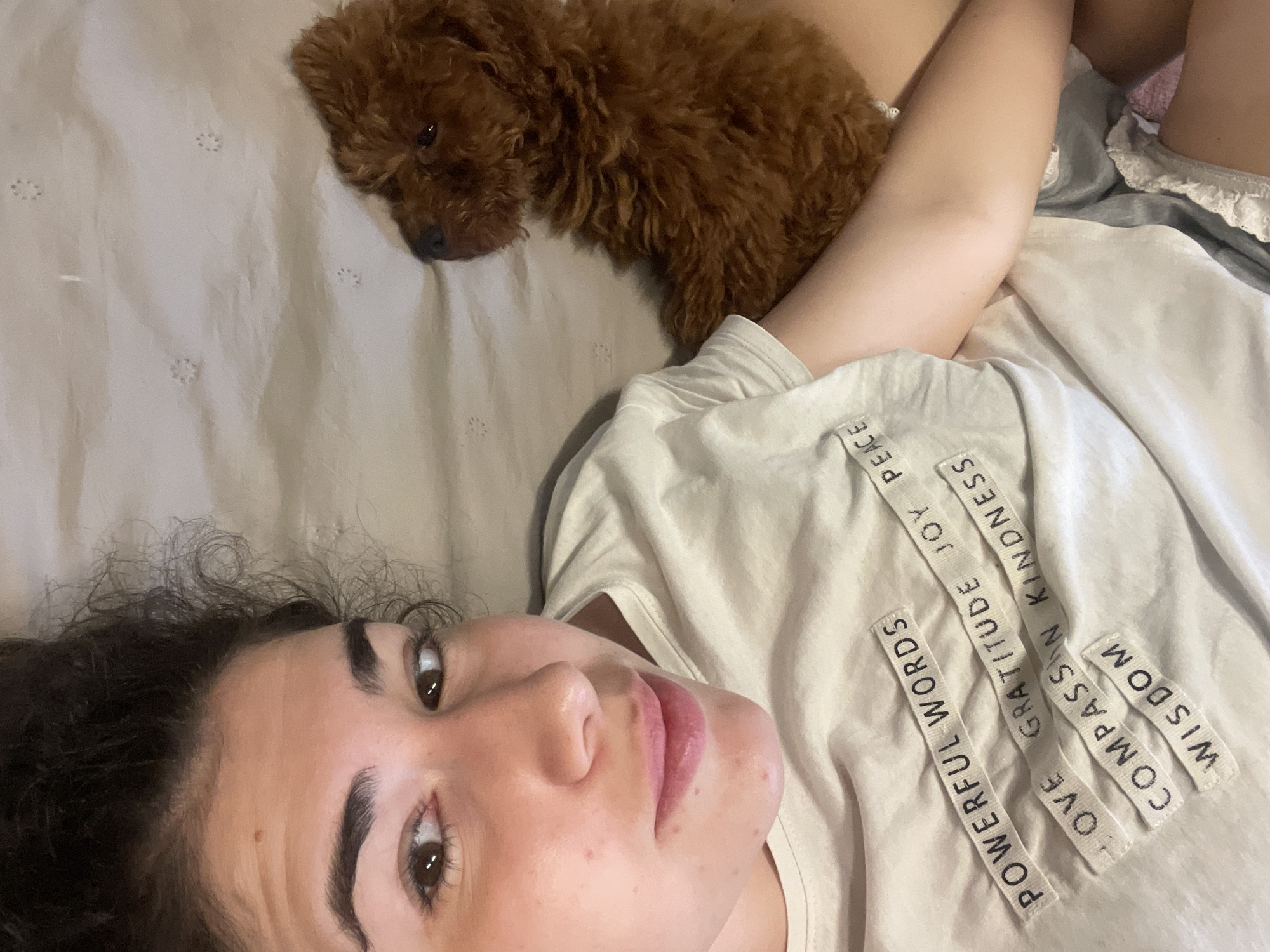 A woman with dark, curly hair is lying on her back on a light-colored bed with a small brown poodle resting on her arm. She is wearing a light-colored t-shirt with words like "POWERFUL WORDS", "GRATITUDE", "KINDNESS", "JOY", "COMPASSION", and "WISDOM" printed on it. The bed has a light-colored sheet with small patterns, and there is another layer of bedding visible. The location is undetermined, and there is no information to suggest it is in Sofia, Bulgaria.