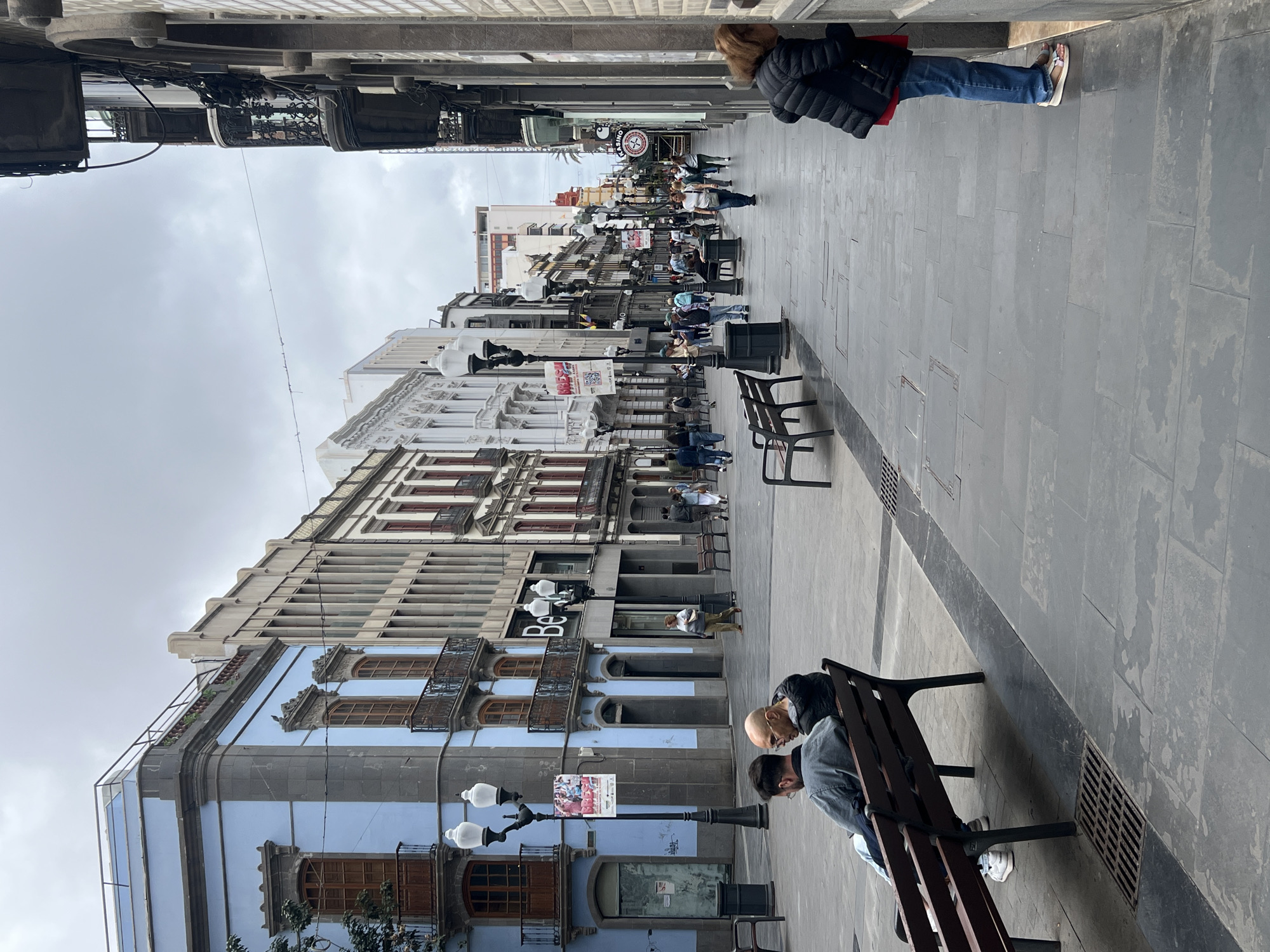 This image depicts a pedestrian street in an urban setting, likely in Las Palmas De Gran Canaria, Spain, under an overcast sky. The street is paved with dark grey square tiles and is lined with multi-story buildings exhibiting traditional European architecture.
In the foreground on the right, a person with blonde hair, dressed in a dark puffer jacket and blue jeans, stands facing away from the camera. Further down the street on the same side, two men are seated on a dark wooden public bench: one is bald with a beard, wearing a dark jacket, and the other has dark hair, wearing a denim jacket. Several other benches are visible along the street.
The buildings on the left side of the street showcase various facades, including one painted light blue with ornate dark balconies and wooden window frames, and another with the partial text "Be" visible, likely part of a store name. Classic-style streetlights with multiple globes are regularly spaced along the thoroughfare.
Many people are visible further down the street, walking, standing, or sitting on benches, indicating a moderately busy scene. In the mid-ground, a prominent round sign with the word "CASINO" is visible above the street level, amidst other commercial signage. The overall atmosphere is that of a typical, albeit cloudy, day in a city center, with people engaged in daily routines.