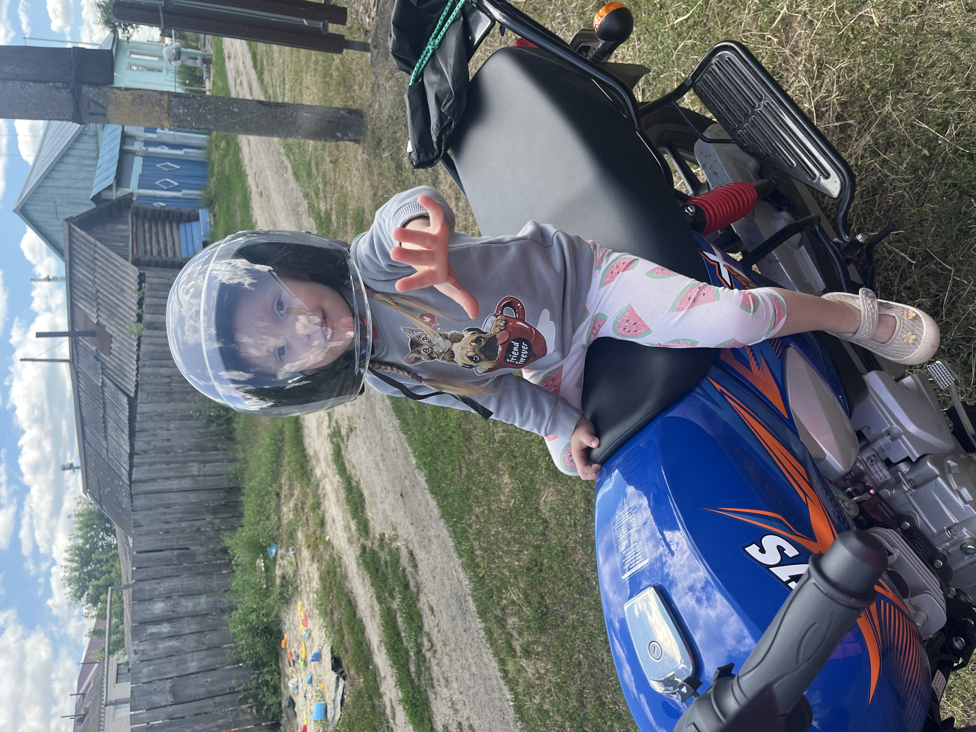 A young child wearing a motorcycle helmet is sitting on a blue and orange motorcycle. The child is wearing a grey sweatshirt with a graphic of two dogs and a mug that reads "Friend forever" and white pants with a watermelon pattern. The child's left hand is raised, with fingers splayed. The motorcycle is parked on a dirt road next to a grassy area. In the background, there are wooden houses and a wooden fence. The sky is blue with white clouds. The image appears to be taken during the daytime on a sunny day. The child's expression is hard to discern due to the helmet. Text visible on the motorcycle includes "SA" and some unreadable white text on a label. The overall scene suggests a rural setting in Сухринское, Russia.