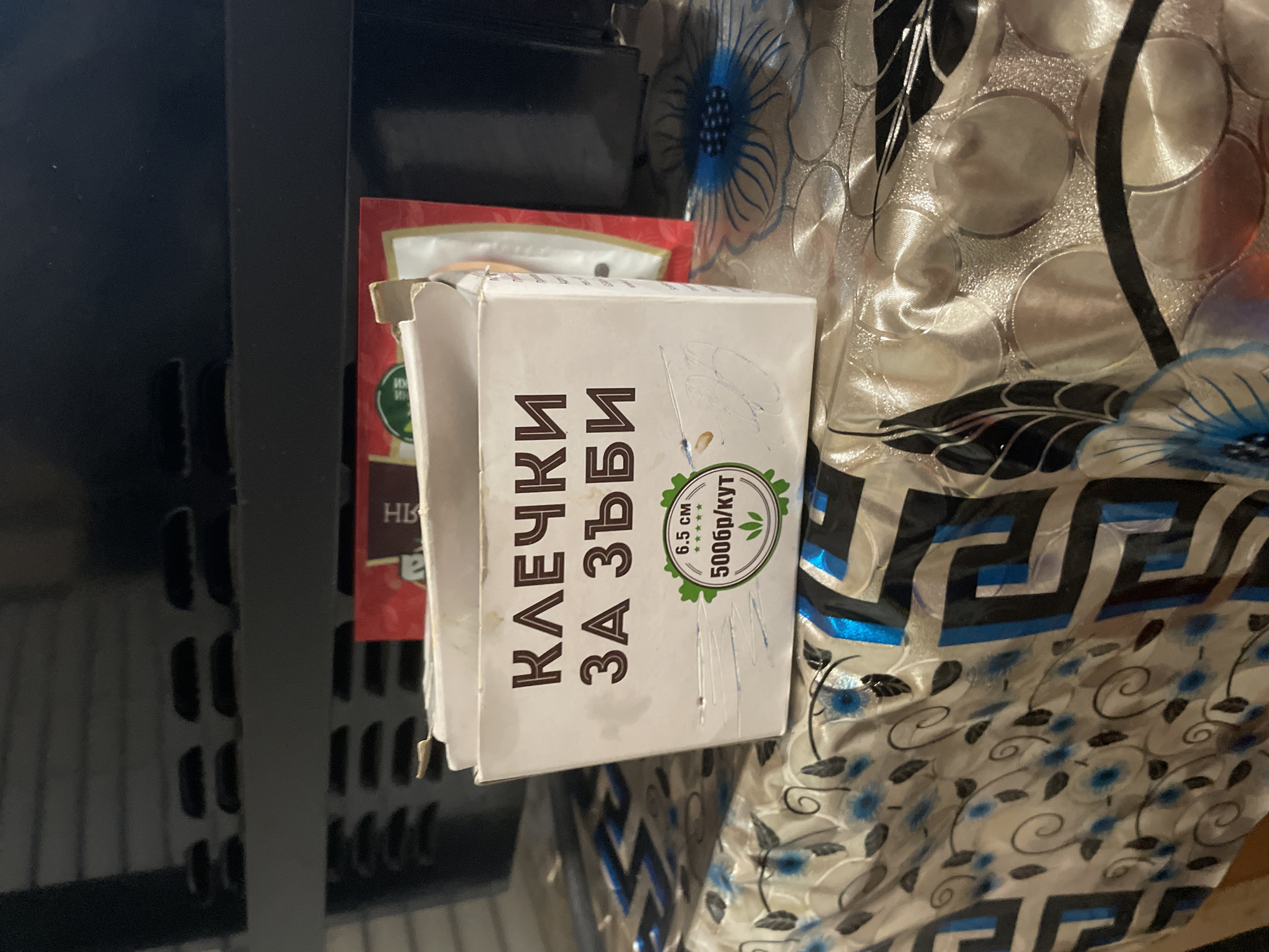 The image displays a close-up, static shot of several objects in an indoor setting, likely in Ihtiman, Bulgaria. The primary subject is a white cardboard box, positioned vertically, featuring Cyrillic text. The most prominent text on the box reads "КЛЕЧКИ ЗА ЗЪБИ," which translates to "TOOTHPICKS." A green circular logo on the box further details the product, stating "6.5 CM" (likely the length of the toothpicks), followed by five stars, and "500бр/КУТ," meaning "500 pieces/BOX."
Behind this white box, a red package is partially visible, showing only the letters "НВ." To the left of the boxes, a dark, possibly black, surface with a slotted or grated design suggests a shelf or part of a furniture item. To the right, a patterned fabric, potentially a tablecloth or decorative cover, is visible. It features a light background with intricate blue floral patterns and abstract black geometric and swirling designs. No people or specific activities are depicted in the scene. The lighting indicates an indoor environment, but the time of day or specific weather conditions are not discernible.