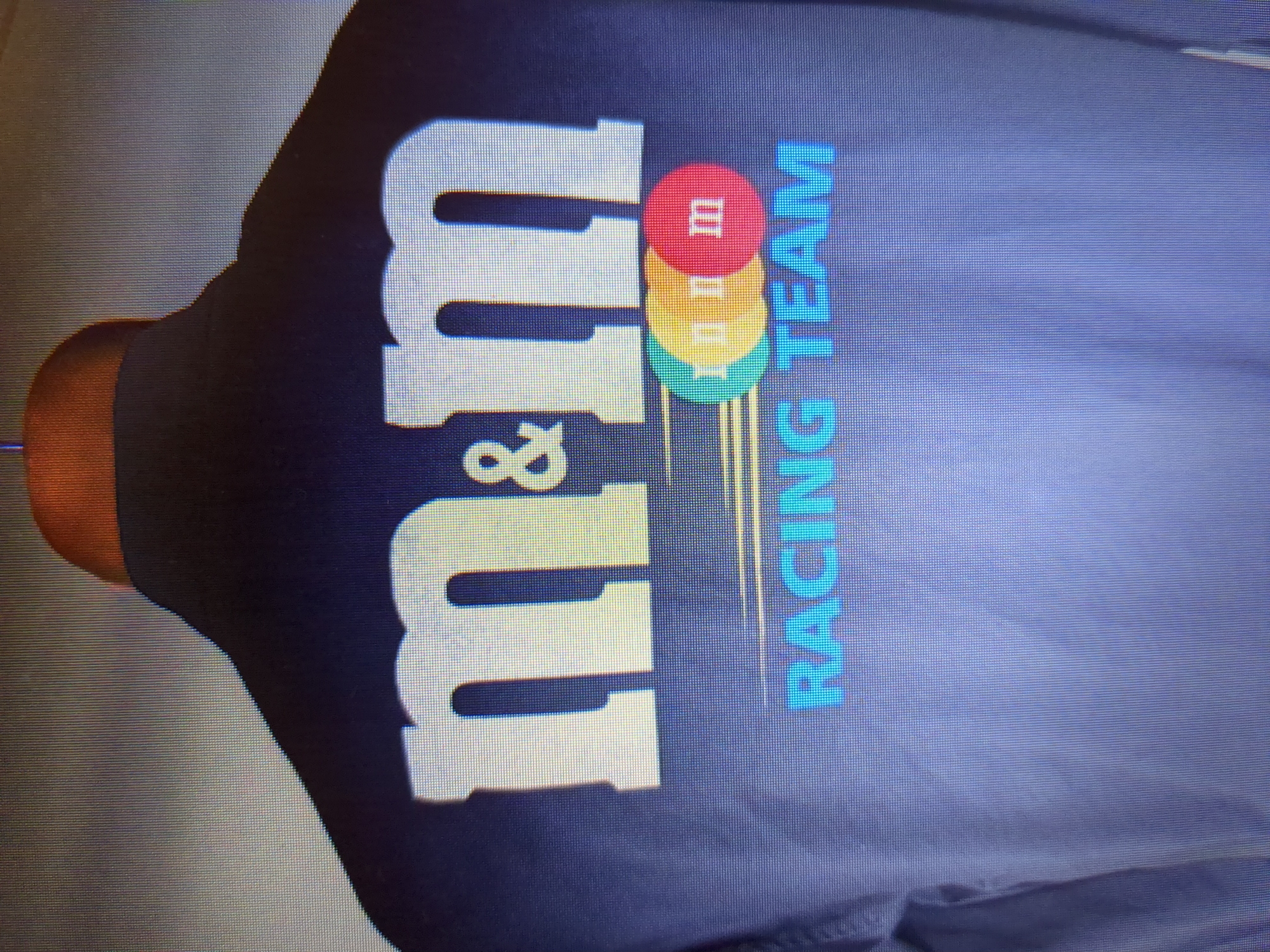 The image shows the back of a dark blue jacket with the M&M's racing team logo. The logo features the text "m&m" in large white lettering, with a smaller ampersand symbol between the two "m"s. Below this, there are three colored circles, representing M&M candies, with the letters "m m m" in white on them. Underneath the circles, the words "RACING TEAM" are written in blue capital letters. The jacket appears to be on a hanger, with the top of the hanger visible at the very top of the image. The background is a gradient of grey and white, suggesting a studio or controlled lighting environment. There are no people or environmental elements visible that would indicate a specific city or location, and therefore no information about Minsk, Belarus, can be extracted from this image.