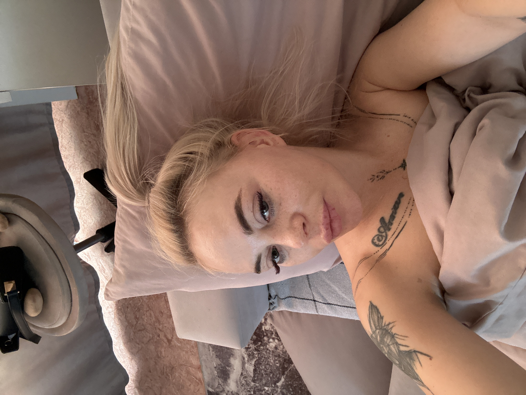 A woman with blonde hair and tattoos is lying on a pink pillow and covered with a pink blanket. The scene is indoors, with a table and other furniture visible in the background. The location is not specified in the image and cannot be inferred.