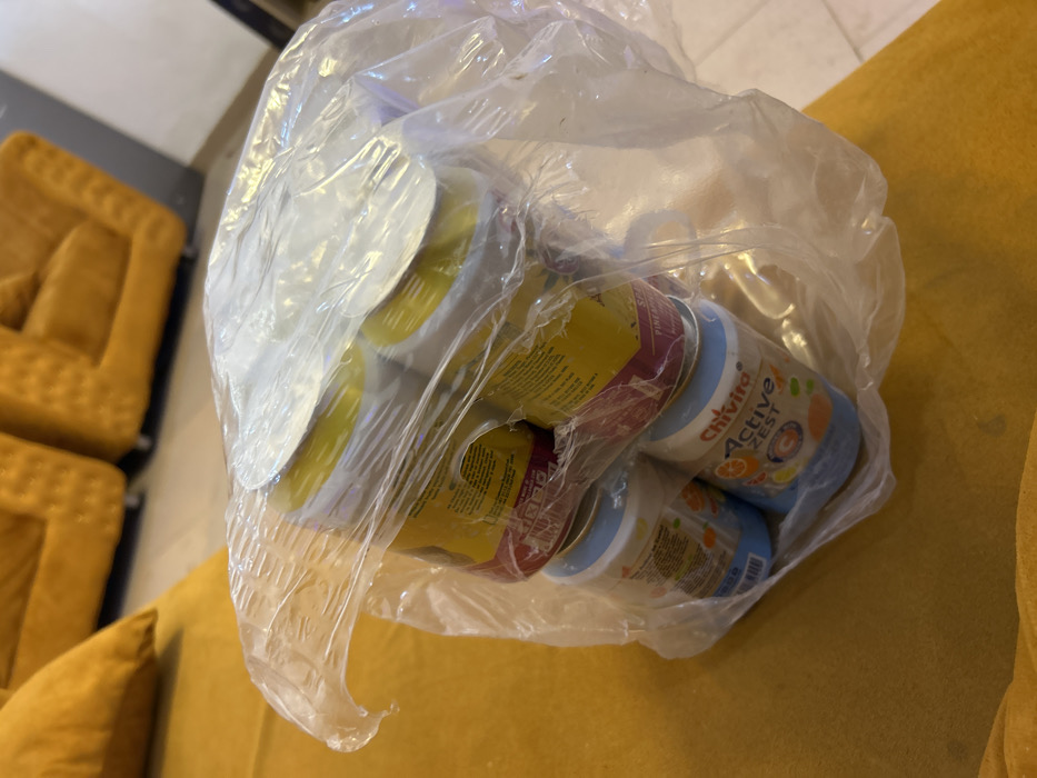 The image displays a clear plastic bag containing six beverage cans, positioned on a yellow upholstered sofa. Four of the cans feature yellow and red labeling, with a partial visible label on one indicating "PINEAPPL." The remaining two cans are distinctly branded "Chivita Active Zest" with blue labels and orange fruit imagery. All cans possess silver foil seals on their tops. The background reveals an indoor environment, characterized by additional yellow/mustard-colored upholstered furniture, including what appears to be a sectional sofa or armchairs. A light-toned floor and a dark wall are also visible in the background. No individuals are present, and no actions or interactions are depicted. The specific location within Egbe, Nigeria, cannot be determined from the visual information provided.