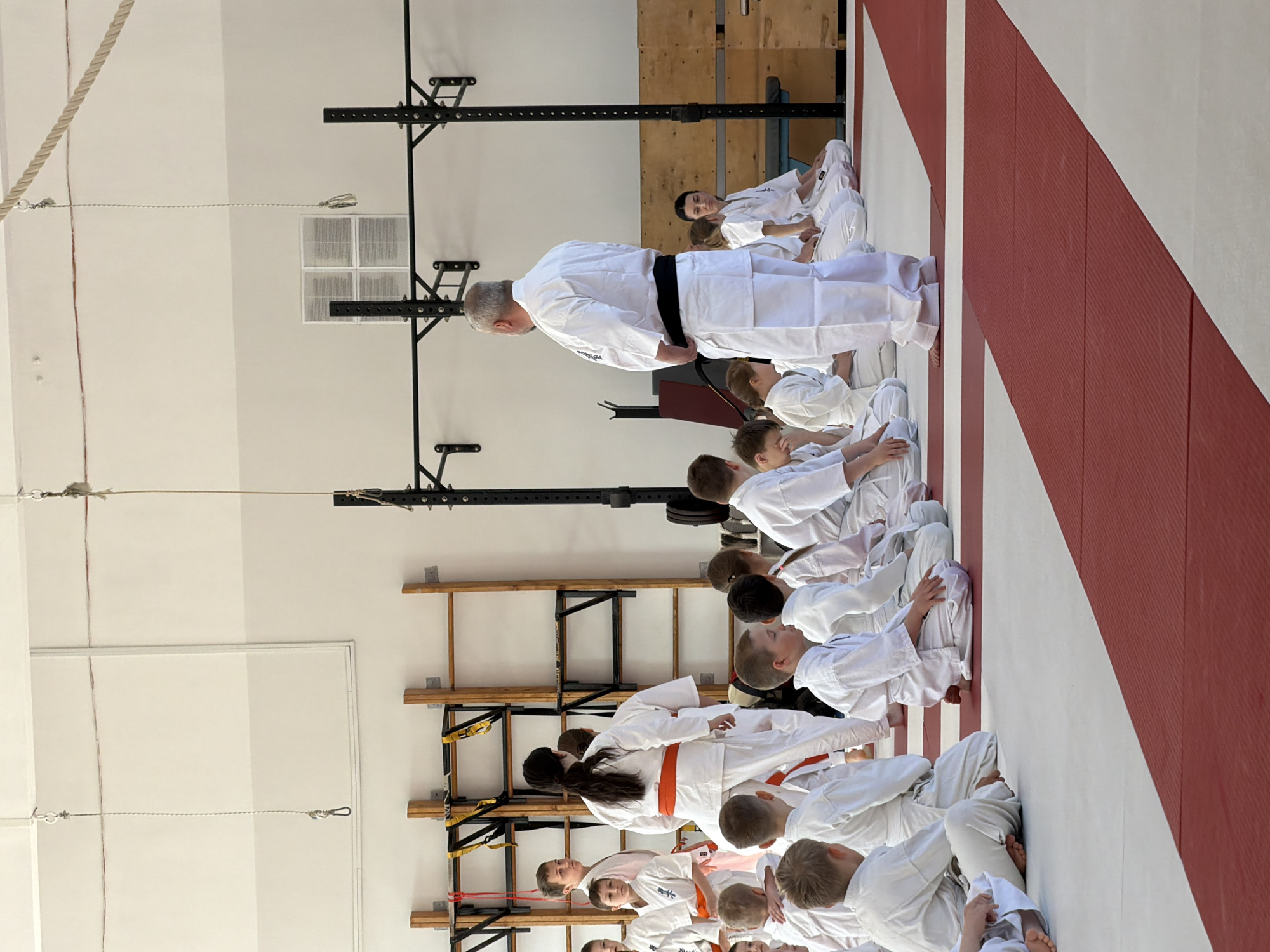 A martial arts class is in session. The scene is indoors, likely a dojo or gymnasium, in what looks like Chaikovsky, Russia. A group of children wearing white martial arts uniforms are kneeling in neat rows. An adult instructor wearing a white uniform and black belt is standing at the head of the rows, facing the children. A girl in an orange belt kneels closer to the front. The room has gym equipment such as parallel bars and a pull-up rig, as well as ropes hanging from the ceiling. Red and white padded walls line one side of the room.