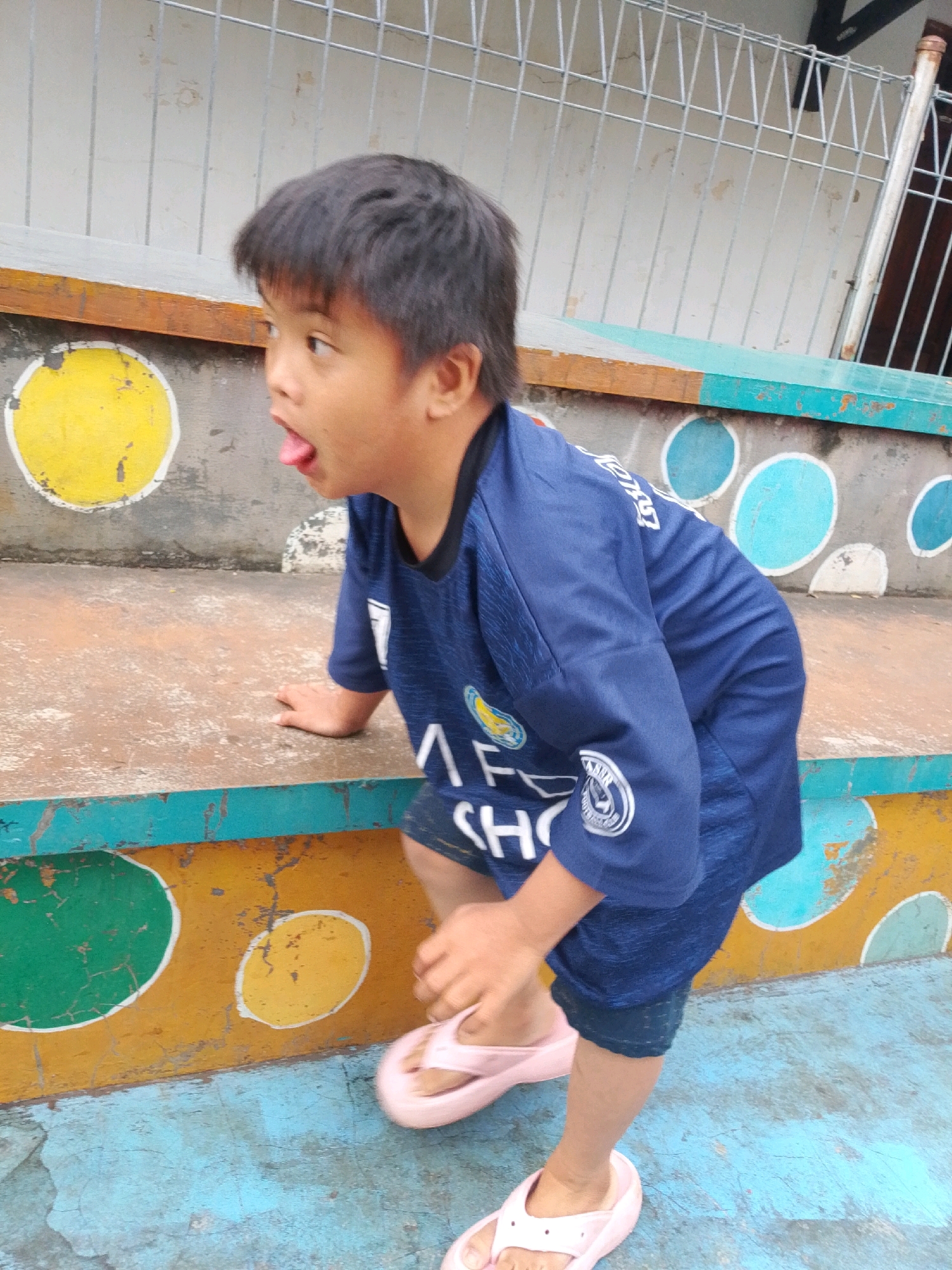 A young boy with dark hair and fair skin is positioned on a weathered, multi-tiered structure, possibly bleachers or steps, painted in vibrant colors and adorned with circles. He is wearing a dark blue jersey with white accents and dark blue shorts, along with light pink flip-flops. The boy is leaning forward, with his left hand on the edge of the structure and his tongue sticking out, as if playing or making a face. The background shows a metal fence and a wall with peeling paint, suggesting an outdoor urban environment. The lighting indicates it is daytime, likely outdoors. The scene appears to be within the city of East Jakarta, Indonesia, given the context provided for the location.