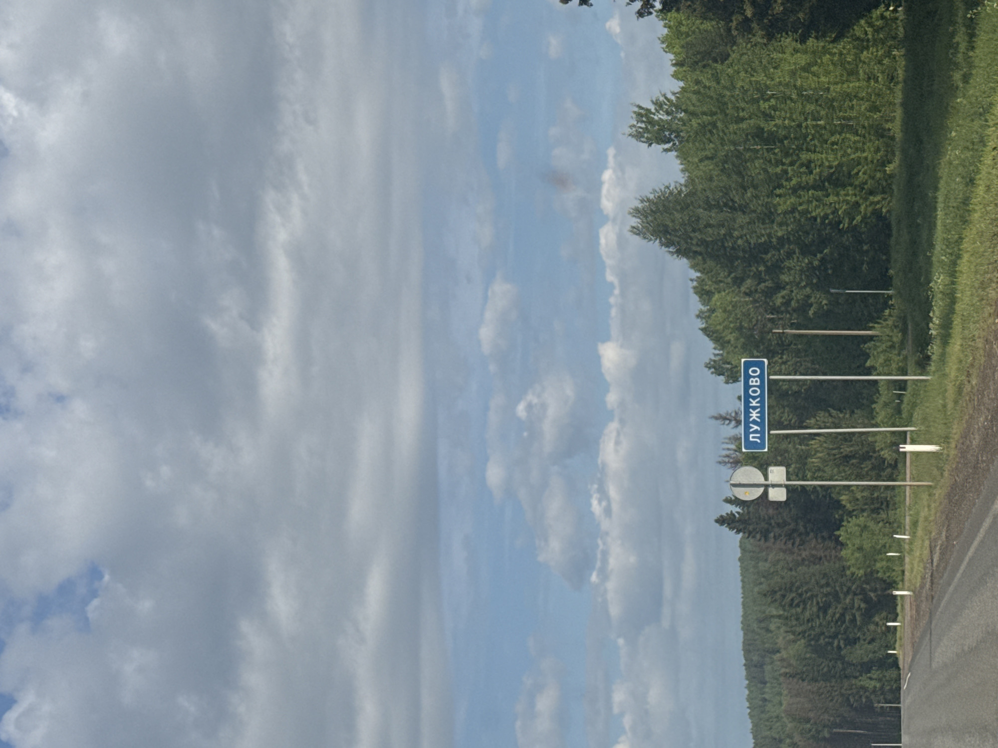 The image displays a rural roadside scene under a partly cloudy sky. A paved road is visible on the right, bordered by a strip of green grass. A dense forest or line of trees runs along the left side of the road, extending into the background. The sky above is bright, with patches of blue visible between scattered white and light grey clouds, indicating daytime.
A blue rectangular road sign with white Cyrillic text is prominently featured on the left, reading "ЛУЖКОВО" (Luzhkovo), identifying the location. Below this, a circular white and grey road sign is visible, likely a traffic regulation sign, possibly indicating "no stopping" or "no parking" due to its shape, though the specific symbol is not clearly discernable. A smaller, indistinct square sign is attached below the circular one. Several white reflective roadside markers line the edge of the road, receding into the distance. No people or vehicles are visible, and the scene conveys a sense of peaceful natural surroundings.