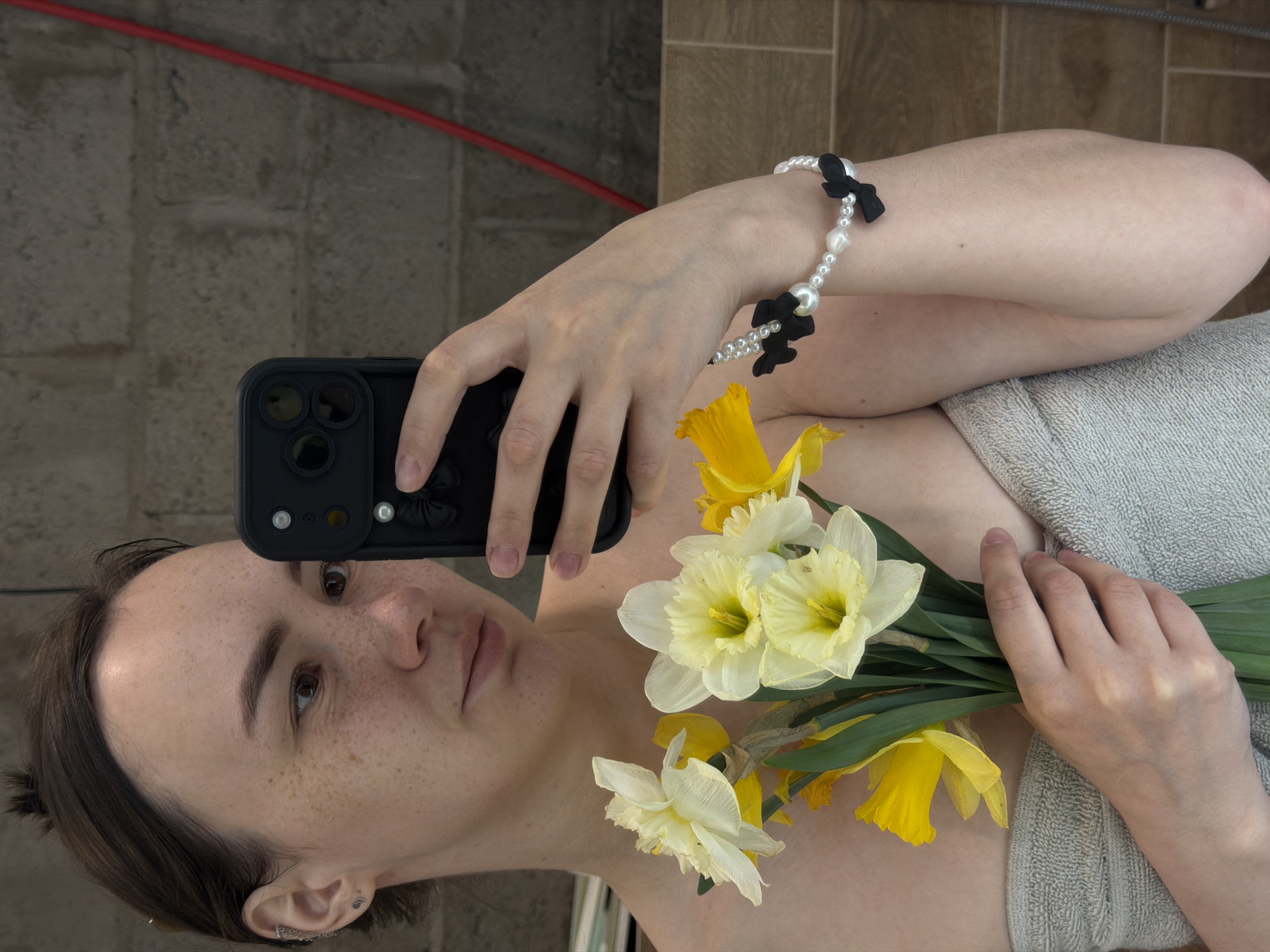 A person lies on their side, facing forward, and holding a black phone in one hand and a bouquet of yellow and white daffodils in the other. They are wearing a pearl bracelet with black bows. The person is partially covered by a gray towel. The background consists of vertically oriented, light-brown tiles, with a red tube visible in the top left corner. The location is not discernible from the image.