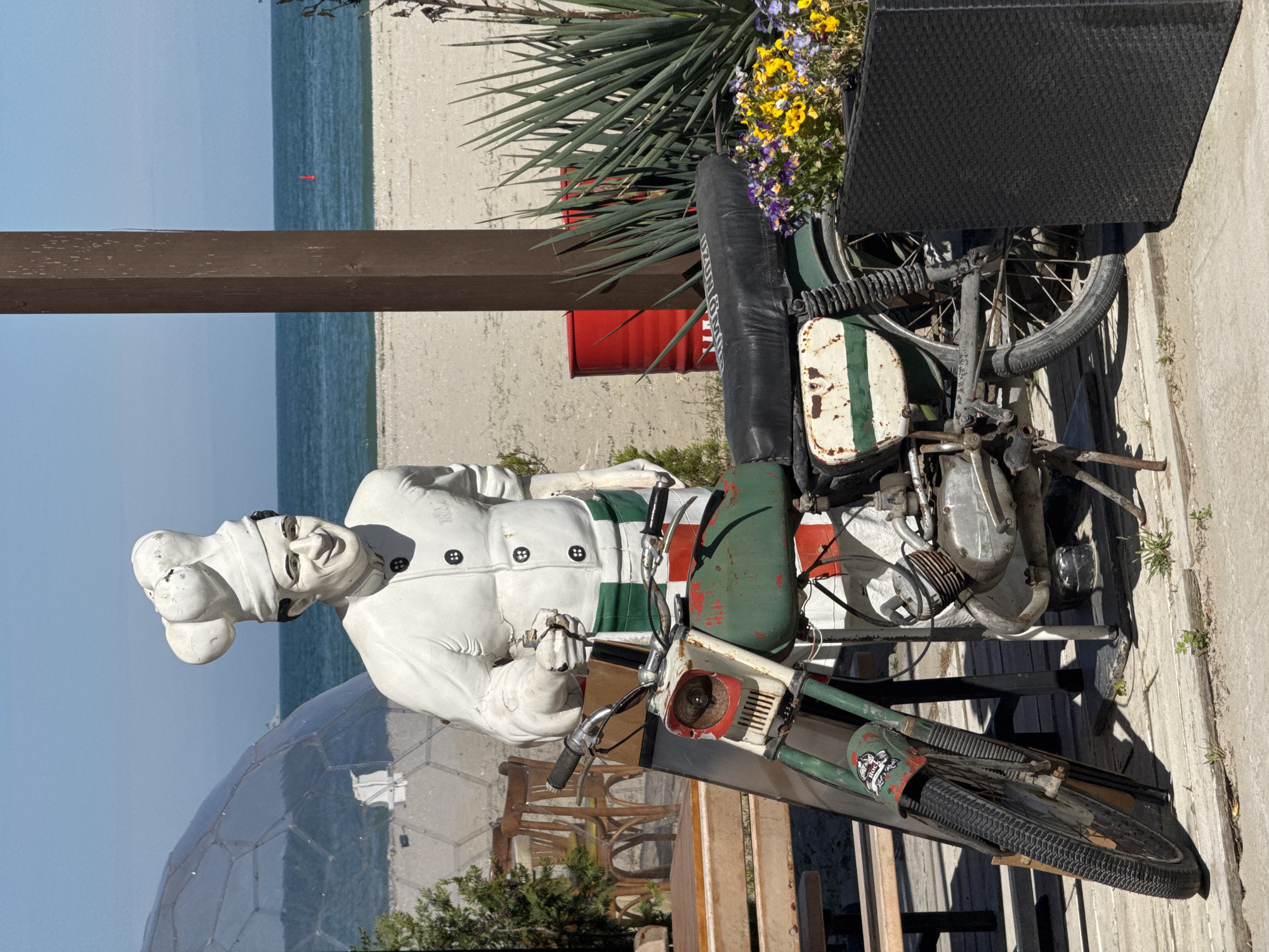 The image shows a statue of a chef sitting on a vintage motorcycle in front of a geodesic dome structure. In the background, there is a beach with sand and water, and a blue sky. The chef is wearing a traditional white chef's hat and uniform. The motorcycle is green, white, and red, and appears to be aged. Flowers are near the rear of the motorcycle. A red barrel is behind the motorcycle. This is likely a decorative display or artistic installation. The location is likely a tourist attraction or restaurant in Varna, Bulgaria, given the chef theme.