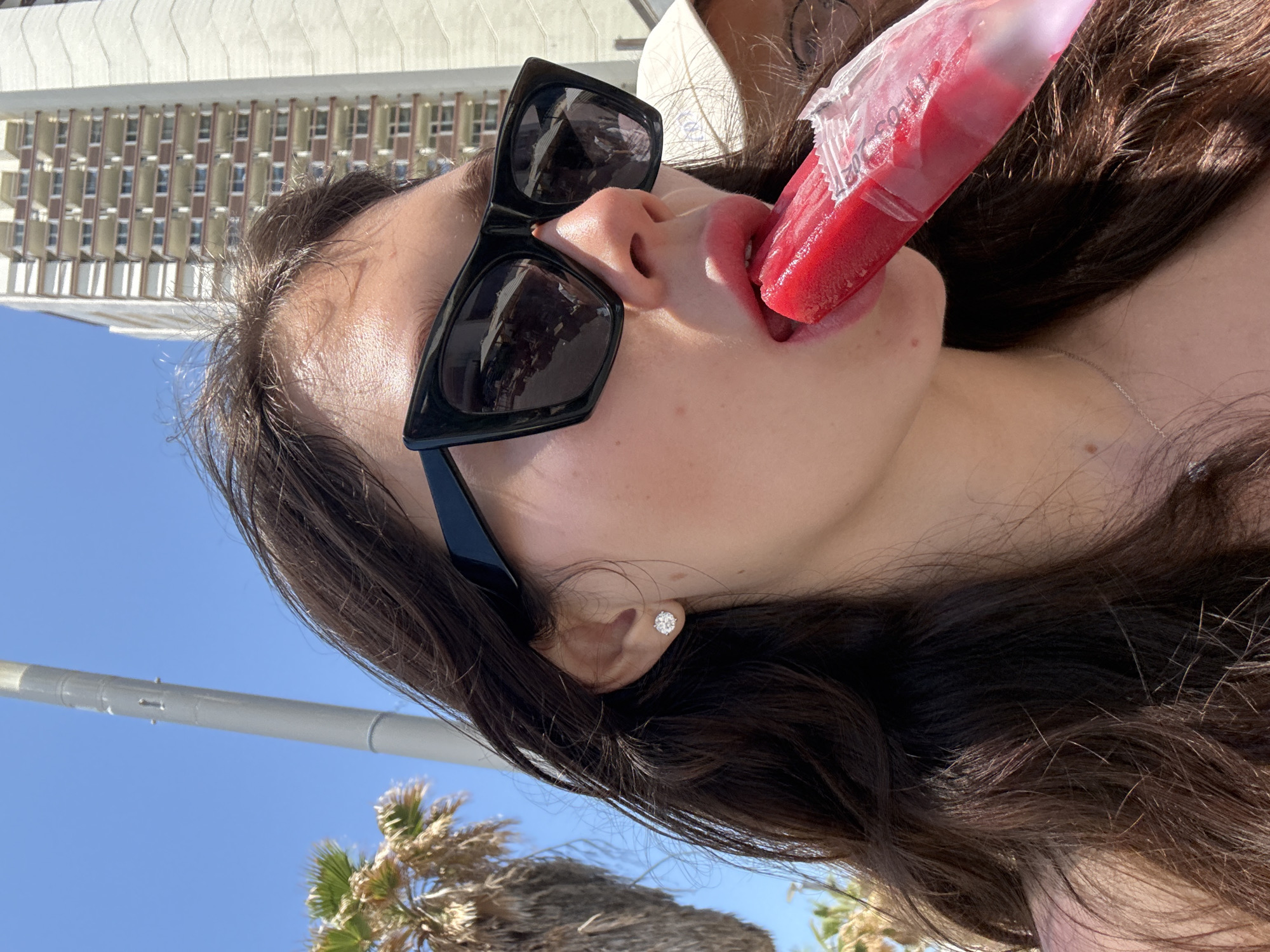 A young woman with dark hair and black sunglasses licks a red popsicle. She is wearing a diamond stud earring in her left ear and a silver necklace. Palm trees are visible in the background. A tall building with multiple windows is also visible. The setting appears to be an outdoor location in Tel-Aviv, Israel, likely near a beach or promenade.