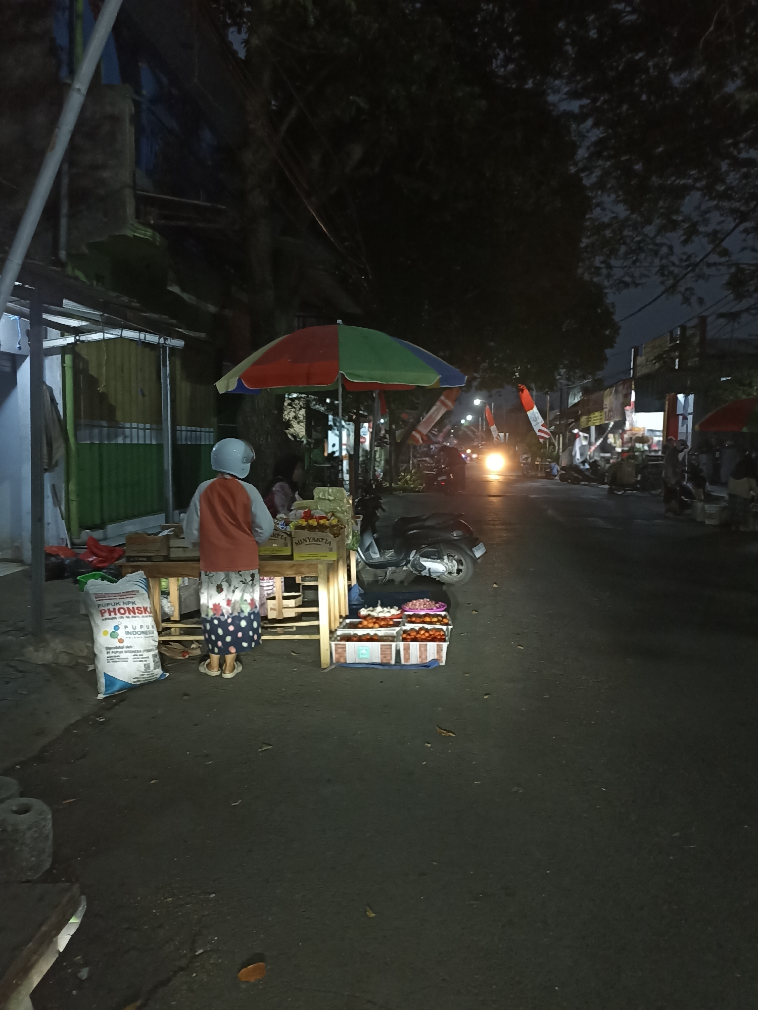 The image depicts a nighttime street scene in Ditotrunan, Indonesia, likely a market area. In the foreground, a person wearing a light blue helmet, an orange and grey shirt, and a patterned skirt stands behind a wooden stall. The stall displays various items, including what appear to be fruits and vegetables in crates, and bags of fertilizer. A large bag labeled "PUPUK NPK PHONSKA" is visible to the left of the stall. A multi-colored umbrella shades the stall. To the right of the stall, a scooter is parked. In the background, traffic lights are visible, and a vehicle's headlights illuminate the street. Indonesian flags are displayed on poles along the roadside. The street is lined with buildings and trees, with lights from various establishments contributing to the ambient illumination.