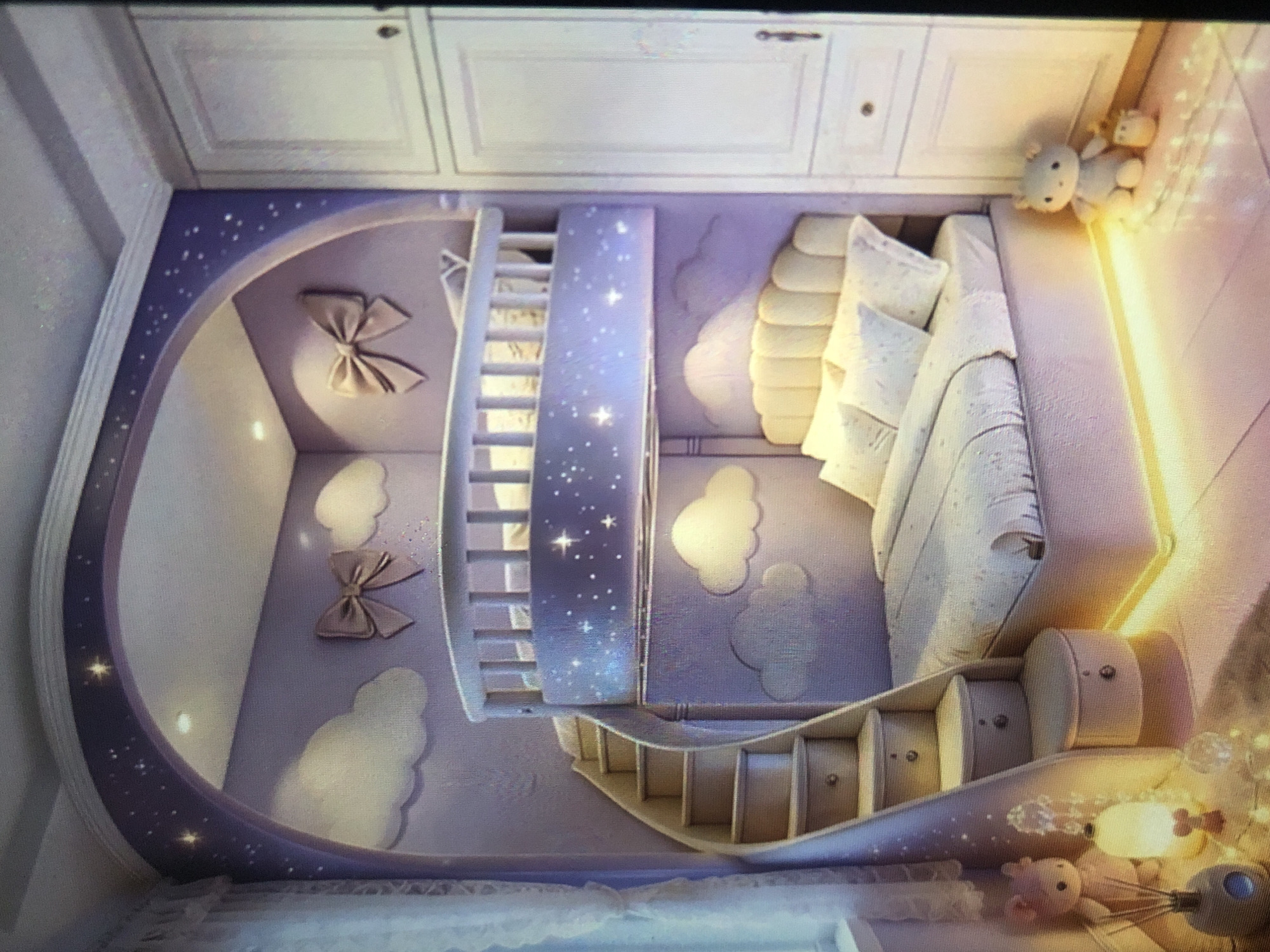The image shows a creatively designed children's room. The walls are painted a light purple with cloud shapes and decorative bows. A bunk bed features a ladder leading to the top bunk, which has starry decorations. The lower section of the room includes a cushioned seating area and a slide leading down from the top bunk. The room has a whimsical, dreamy aesthetic. Two stuffed animal toys are situated around the seating area, and another one on the bunkbed. A set of white cabinets is mounted above the room. There is no information to determine whether this is in the city of Lhokseumawe, Indonesia.