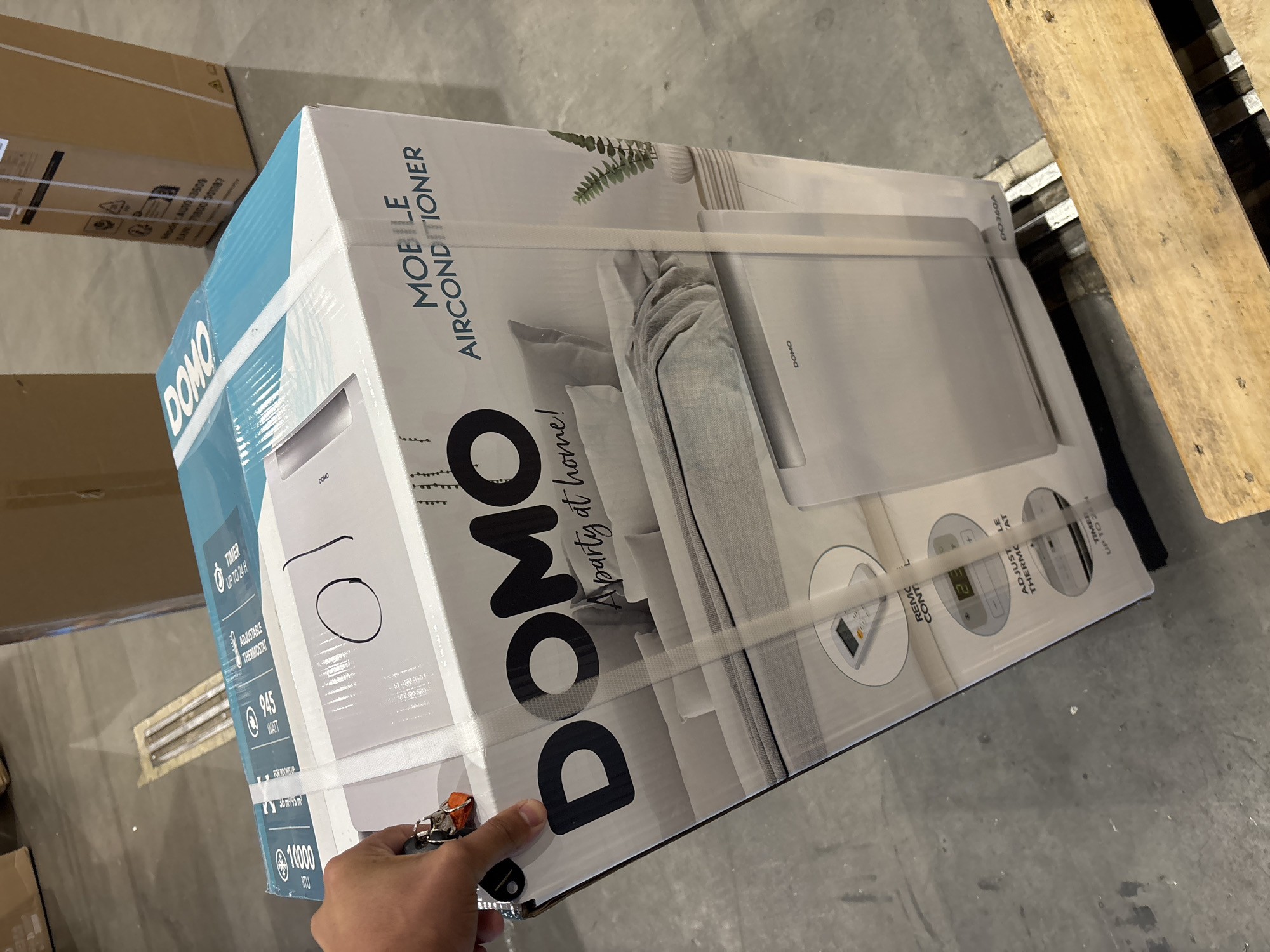 An overhead medium shot shows a white and turquoise cardboard box for a "DOMO MOBILE AIRCONDITIONER" positioned on a wooden pallet. The box is oriented vertically, with the front facing the camera. The brand name "DOMO" is prominently displayed in large black lettering. Below this, text reads "Apartment at home!". The lower portion of the box features an illustration of a bedroom scene with a bed and a white air conditioning unit. Various circular icons detailing features like "REMOTE CONTROL", "ADJUSTABLE THERMOSTAT", and "TIMER UP TO 24H" are visible. A person's hand, holding a keyring with several keys, is reaching from the left side of the frame, partially obscuring the top left corner of the box. The background consists of a concrete floor with other cardboard boxes and industrial elements, suggesting a warehouse or storage environment. The scene appears to be indoors, with artificial lighting. The presence of the wooden pallet indicates the item is likely being stored or prepared for transport.