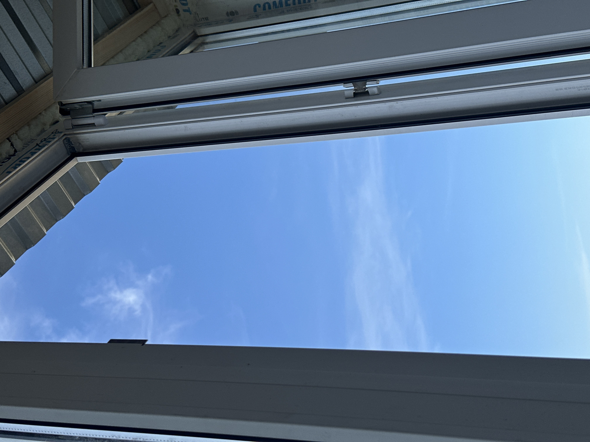 The image shows a view of the sky through an open window frame. The window appears to be made of white PVC and is partially open. The sky is a clear blue with a few wispy white clouds. In the background, there is a glimpse of a corrugated metal roof and some wooden beams, suggesting that this is an interior view from a building under construction or renovation. There are no people or any specific landmarks visible that would indicate the location within the city of Energetik, Russia. The image is a close-up shot, focusing on the window and the sky.