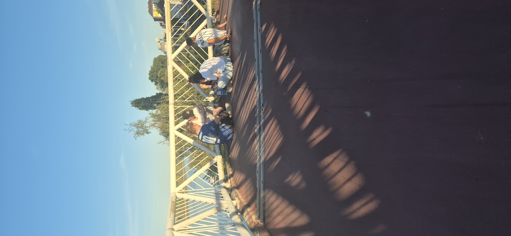 Four individuals are seated on the ground of a pedestrian walkway on a bridge in Mainz, Germany, facing a body of water. The individual on the far left is wearing a blue soccer jersey with the number 10. The person next to them, to the right, is wearing a black baseball cap and a white shirt. The third person from the left is wearing a white t-shirt and jeans, and the fourth person is wearing a white t-shirt and dark pants, facing away from the camera. All four individuals appear to be engaged with their mobile phones. A white metal railing with a diamond pattern runs along the edge of the bridge, separating the walkway from the water. The sky is a clear blue, and trees and buildings are visible in the background. Shadows from the railing are cast across the walkway.