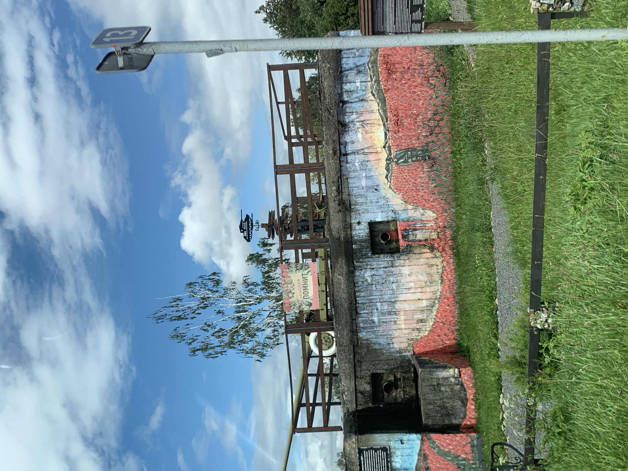 The image shows a concrete fortification, likely a bunker, with a large cannon barrel protruding from the left side. The bunker is painted with a mural depicting a field of red flowers and a figure standing in them, under a sky with clouds. A sign with Russian text "Беларусь помнит" (Belarus Remembers) is attached to the railing on top of the bunker. There is a wooden fence in the foreground, with green grass and some gravel paths. In the background, there are trees and a signpost with a number "13" and other directional signs. The setting appears to be an outdoor historical memorial or museum in Цімкавіцкі сельскі Савет, Belarus.