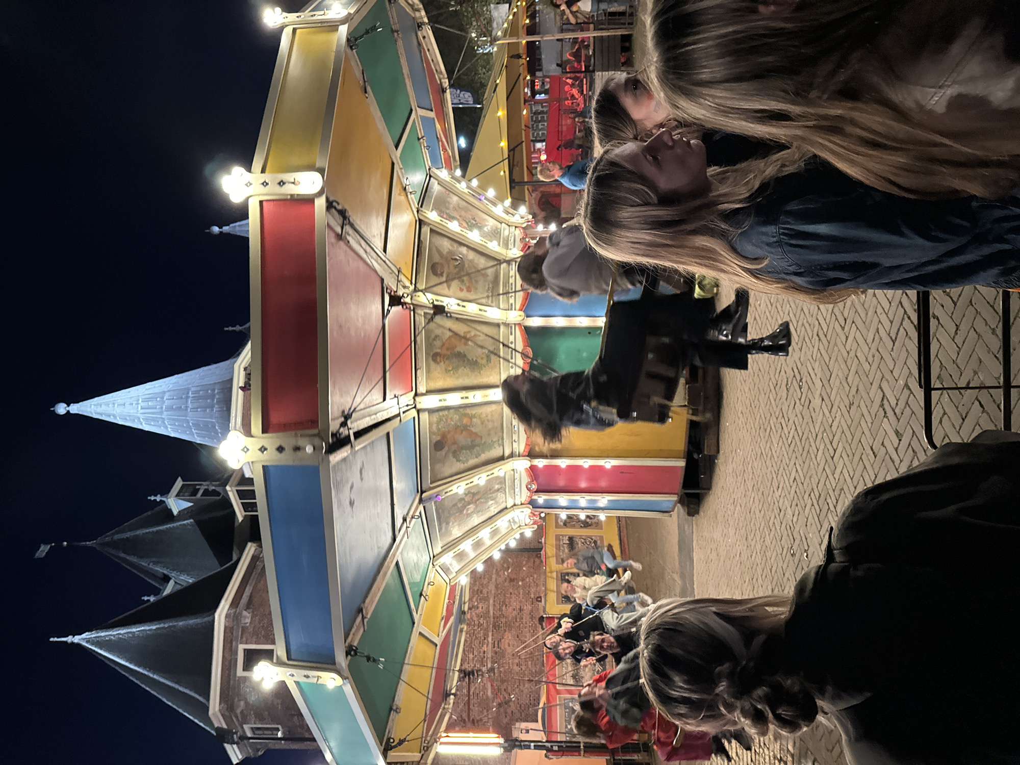A carousel amusement ride in Amsterdam, Netherlands, is shown in operation at night. The carousel has a series of seats that are suspended on chains that people are riding on. The carousel is painted in a variety of colors, including red, yellow, green, and blue, with white lights hanging from the roof. In the background, there is a dark sky. People are standing in the foreground, looking at the carousel, their faces and hair are illuminated by the light from the carousel. The ground is paved with brick.