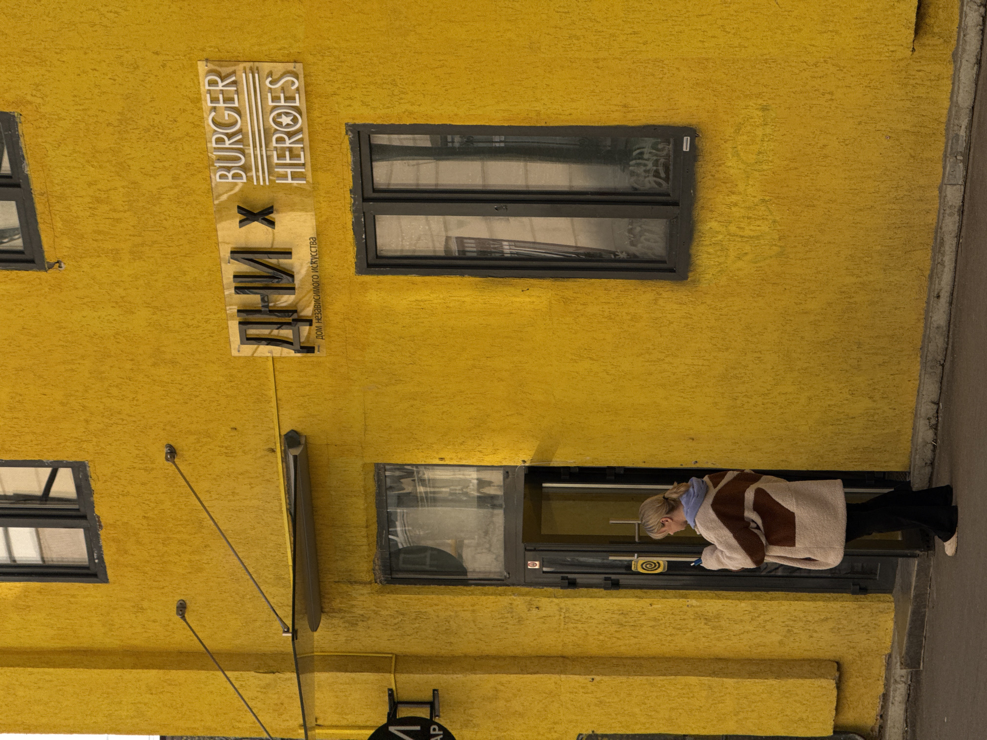 A vertical shot depicts a yellow building in Moscow, Russia. The sign "Burger Heroes" is seen at the top. Below it is the sign "ДНИ Х", which translates to "Days X," with the subtitle "дом независимого искусства" meaning "house of independent art." The building features dark gray windows with graffiti visible through them, and a woman is standing in the doorway, looking at her phone. She is wearing a beige and brown patterned jacket, black pants, and white shoes. The environment is urban, and the building’s texture appears rough.