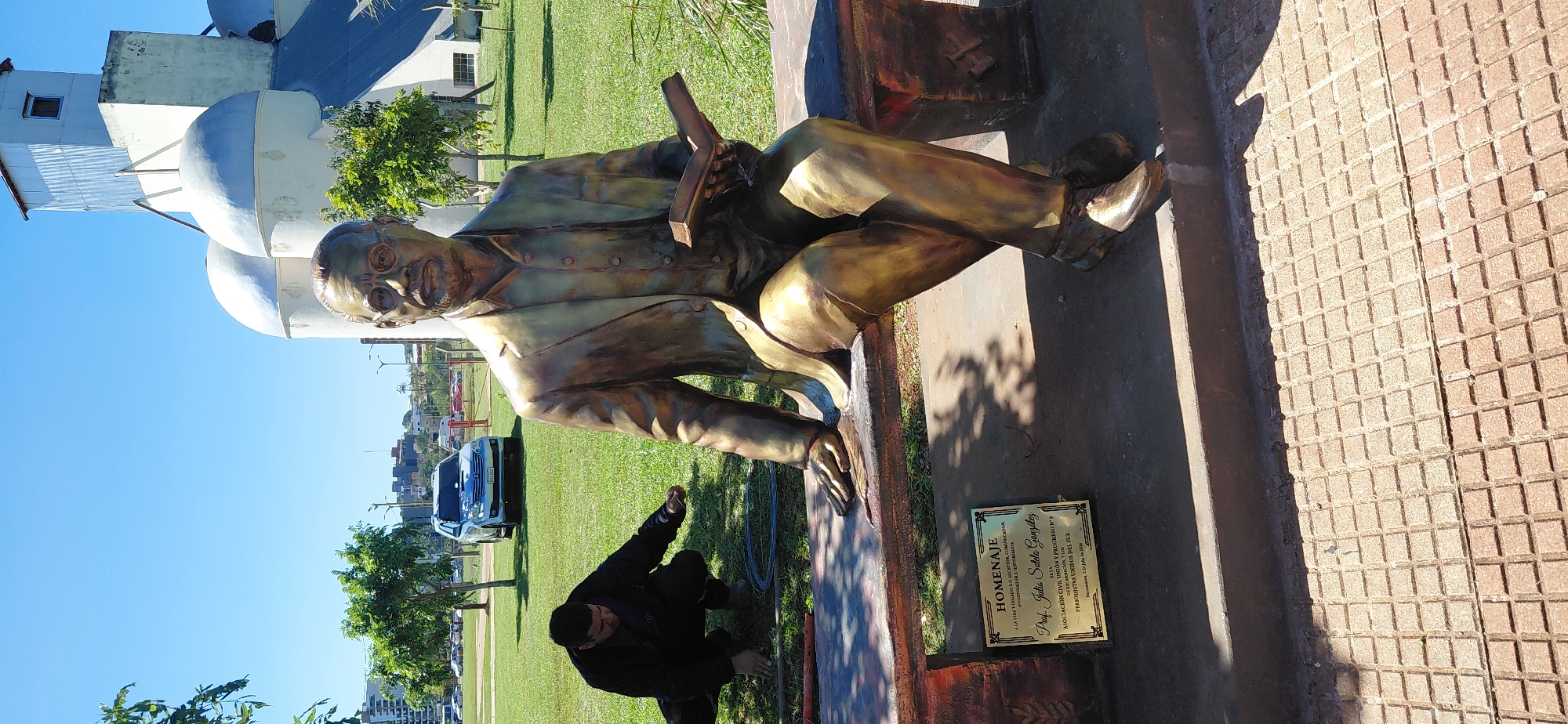 The image depicts a bronze statue of a man sitting on a bench, holding a book. The statue is located in a park with a grassy area, trees, and a clear blue sky. In the background, there are some buildings, including silos and a car parked on a road. A man is crouching on the ground in front of the bench, appearing to be working on something. A plaque is visible on the bench, inscribed with "HOMENAJE" and other text. The statue and plaque are in a public outdoor space, likely a monument or memorial within the city of Encarnacion, Paraguay.