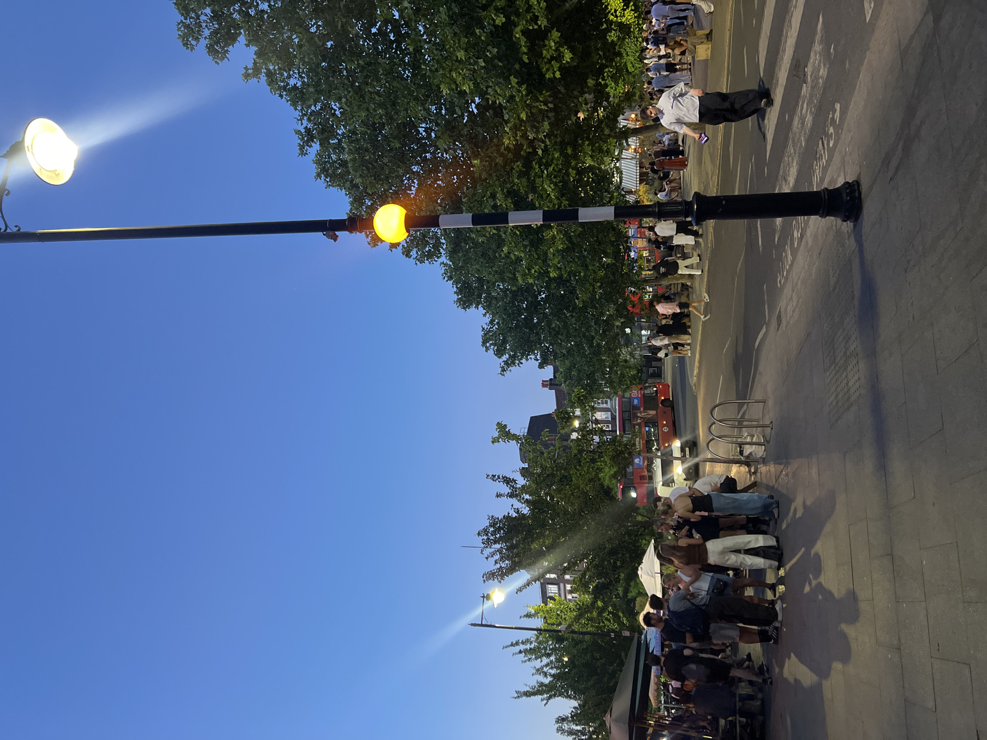The image shows a street scene in London, United Kingdom. A street lamp is in the foreground, with a clear blue sky above. A pedestrian walkway or sidewalk borders the street. Groups of people are gathered along the sidewalk, likely engaged in socializing or waiting for something. In the background, a red double-decker bus is visible, a common sight in London. Buildings are also visible in the background, obscured partially by trees.