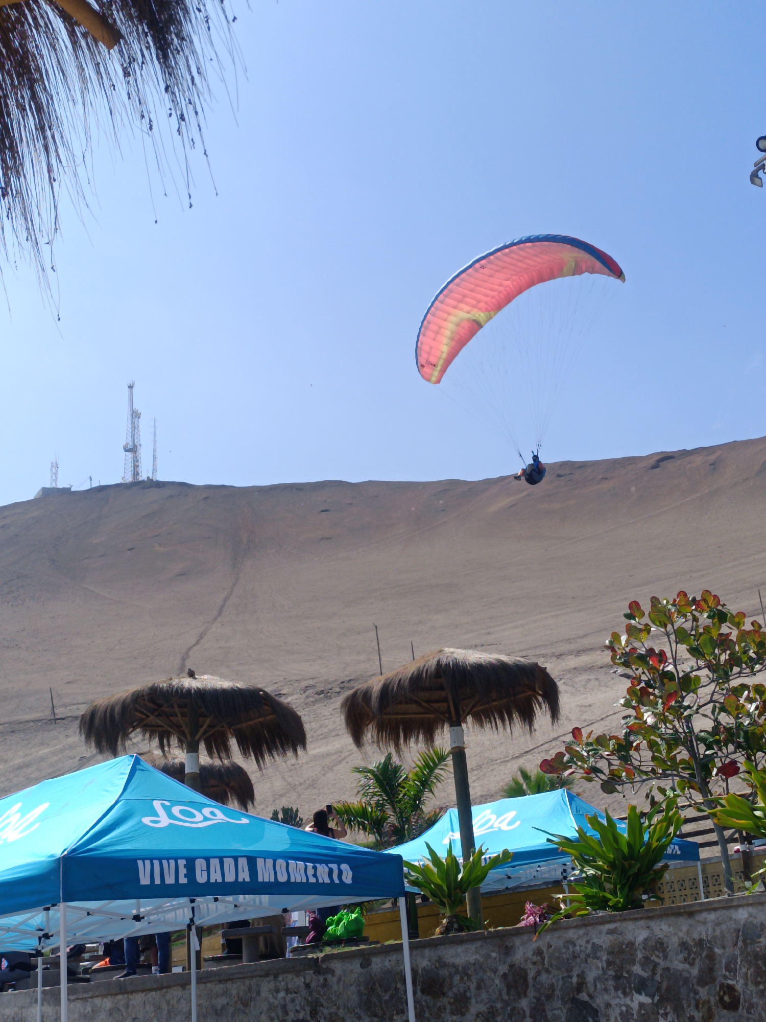 This image depicts a sunny day at a coastal location in Arica, Chile. In the sky, a paraglider with a red and yellow parachute is soaring, with a person suspended beneath it. In the background, a barren, sandy hill rises, topped with several communication towers. In the foreground, two blue tents with white lettering that reads "VIVE CADA MOMENTO" are set up, suggesting a recreational or event area. Several thatched umbrellas are visible behind the tents, offering shade. A person is seen taking a photograph in the background, and some foliage and a stone wall are present in the lower part of the image. The overall scene suggests a popular outdoor activity spot.