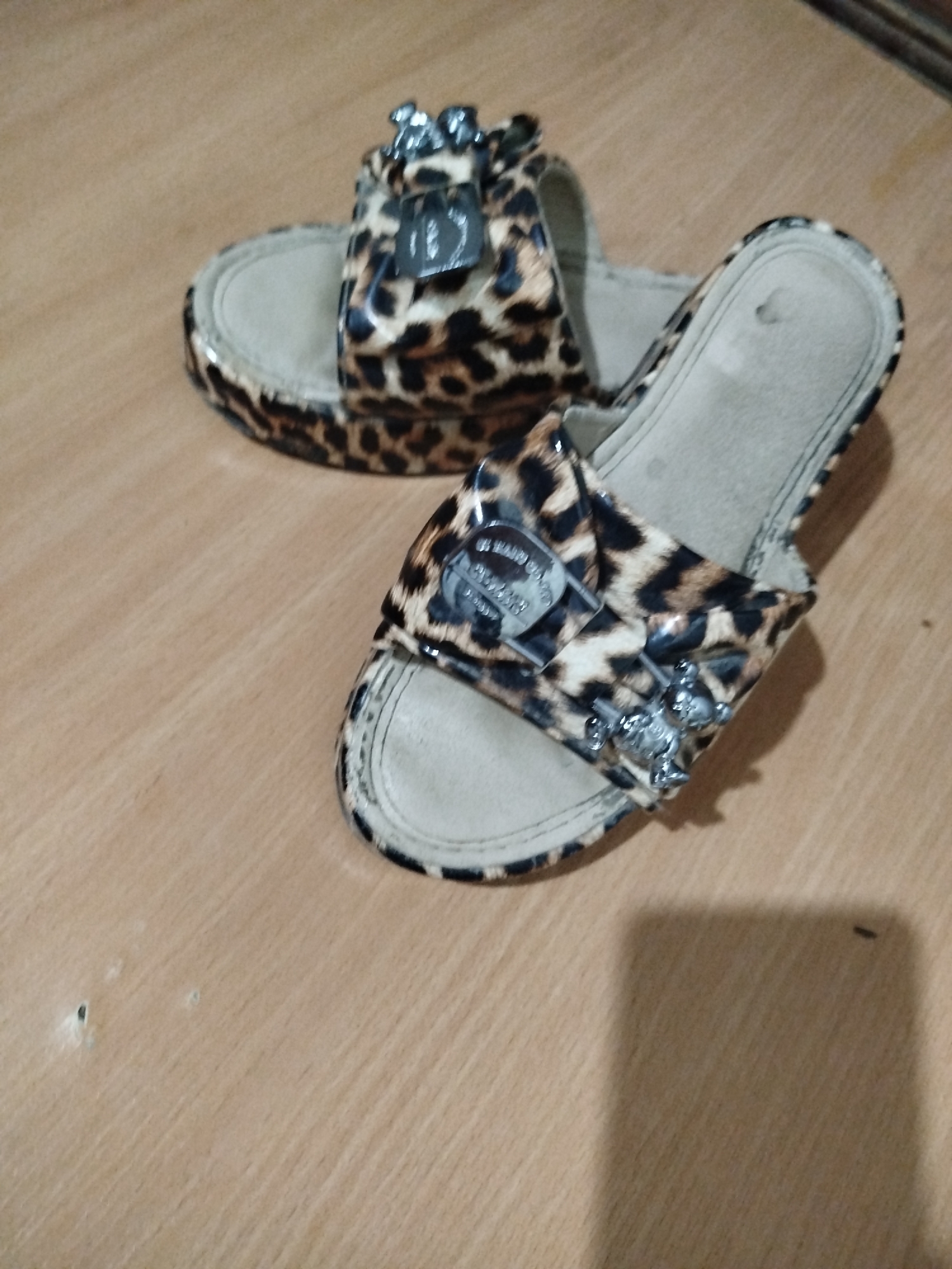 A pair of leopard-print slide sandals with silver embellishments is displayed on a wooden surface. The sandals appear to be women's footwear, with a wide strap across the top of each foot. The straps are adorned with what look like small lock charms and some sort of shiny decorative elements. The soles of the sandals are a light tan color. The wooden surface is a light brown with visible grain. The lighting in the image suggests an indoor environment, possibly a home. There are a few small, dark specks on the wooden surface. The scene is static, with no visible human interaction or action. The location is not specified beyond being in the city of Unknown, Unknown.