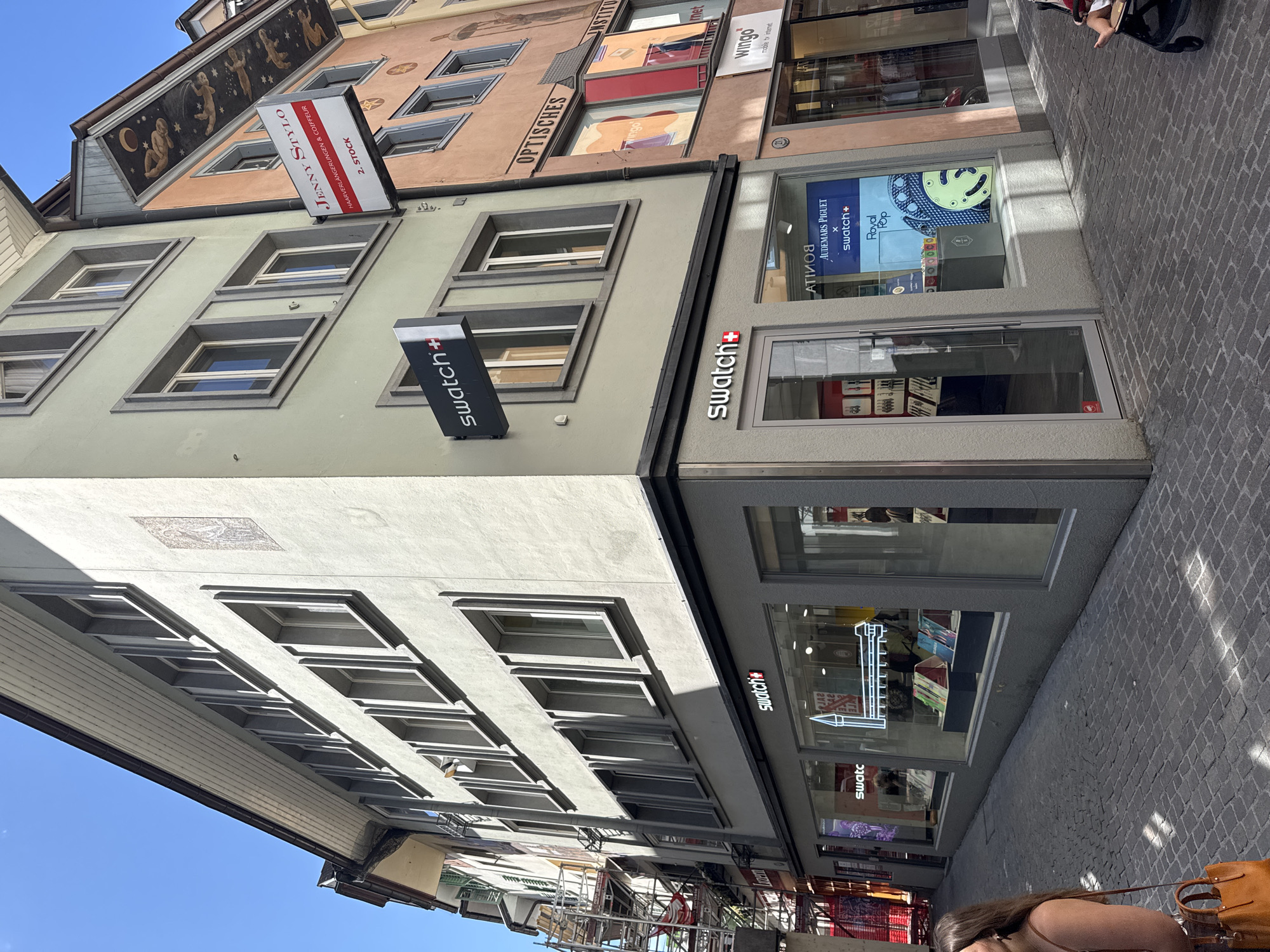 The image shows a street view in Lucerne, Switzerland, focusing on the corner of a building housing a Swatch store. The Swatch store has a prominent storefront with large glass windows displaying merchandise and promotional materials. Above the store, a Swatch sign is visible on the building's facade. Adjacent to the Swatch store is another building with a sign for "JENNY STYLO HAARVERLANGERUNGEN & COIFFEUR" on its second floor. The buildings are typical of European urban architecture, with multiple windows and decorative elements. The street is paved with cobblestones, and sunlight casts shadows on the ground. In the foreground, part of a woman carrying a brown handbag is visible on the left. On the right, a person is seated in a stroller. The overall scene depicts a commercial street in a city, likely a tourist area, given the presence of well-known brands like Swatch.
