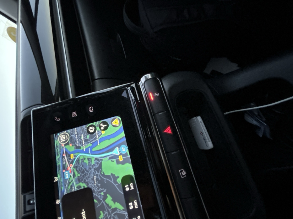 The car's infotainment system displays a navigation map with a route highlighted in blue, showing a distance of 61 km and an estimated time of 54 minutes. The map includes details of roads, green areas, and points of interest. The center console features buttons, one with a red lock symbol illuminated and another with a red triangle for hazard lights. A small white object, possibly a USB drive, is visible in a storage compartment.