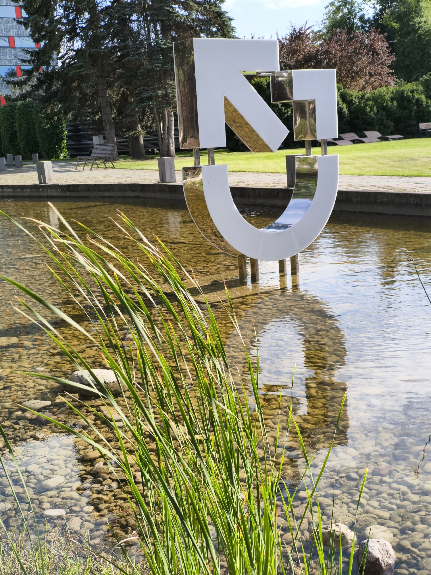The image captures a modern art sculpture featuring a large white structure with metallic accents, positioned over a shallow pond filled with clear water and rounded pebbles. The sculpture is partially submerged in the pond, with its reflection visible on the water's surface. Tall green reeds grow along the edge of the pond in the foreground, obscuring some of the lower parts of the sculpture. In the background, there is a well-maintained lawn, several trees with dark green and reddish foliage, and some lounge chairs. A modern building with a facade featuring red and white rectangular elements is visible to the left of the frame, partially obscured by trees. The sky is blue with scattered white clouds. The scene appears to be an outdoor public space, possibly a park or the grounds of an institution in the city of Tallinn, Estonia. There are no people or animals visible in the image.