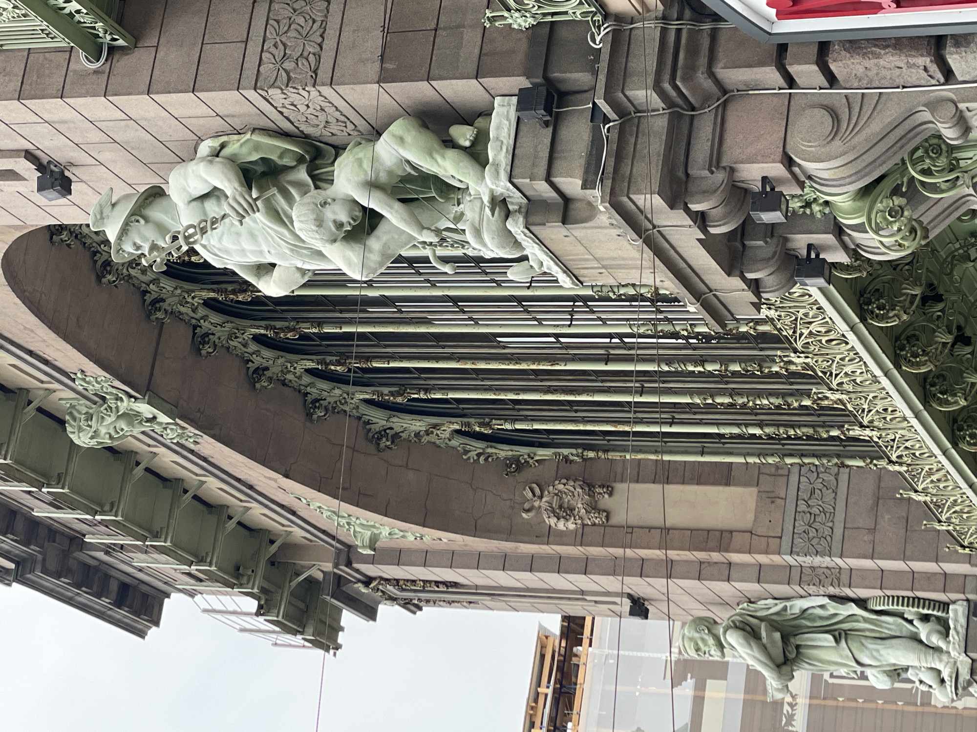 The image shows a low-angle view of architectural details on a building. Three sculpted figures, possibly male nudes, are visible near the top of the frame, likely part of a cornice or decorative feature. The sculptures appear to be made of light-colored stone or plaster. The architecture around the sculptures has a grey-green color with intricate metalwork and stone carvings. The setting seems to be a historic or ornate building, as indicated by the level of architectural detail. The location is Saint Petersburg, Russia, specifically the facade of the Eliseev Emporium.