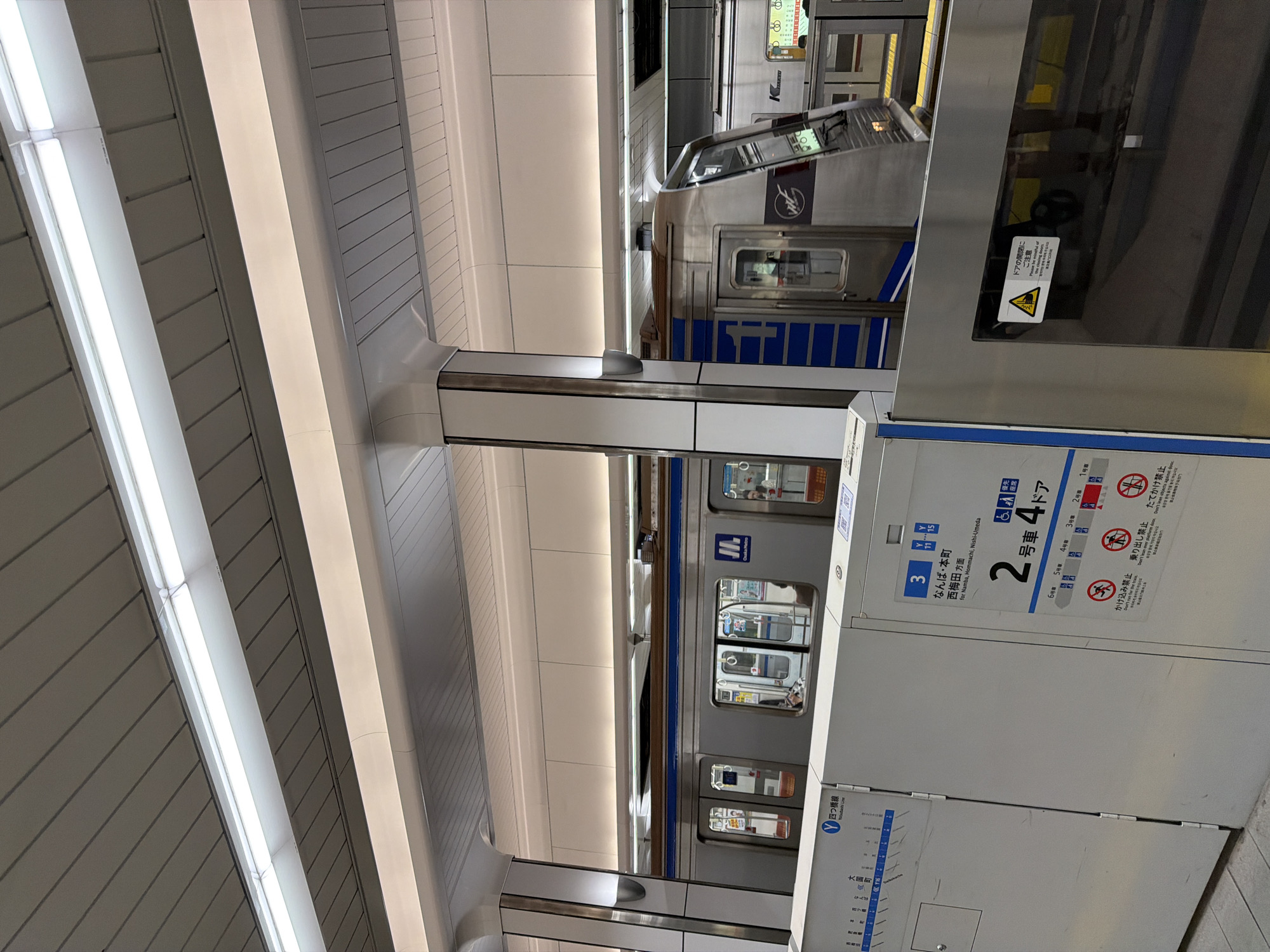 An indoor scene depicts a modern subway or train station platform, likely in Osaka, Japan, based on visible text. A silver train with blue accents is stopped at the platform, its doors closed. Reflections are visible on the train's windows and metallic surfaces of the platform safety barriers.
The platform features sleek, white and grey panelling on the walls and ceiling, illuminated by long, bright fluorescent light strips. A robust support pillar stands between the platform edge and the barrier. Informational signage is mounted on the platform-side barriers, providing guidance to passengers.
Visible text on the signs includes "四つ橋線" (Yotsubashi Line), indicating the specific train line. Directions for "なんば・本町 西梅田方面" (for Namba, Hommachi, Nishi-Umeda) are displayed, along with a destination map showing "大国町" (Daikokuchō). A specific car number and door indication "2号車 4ドア" (Car No. 2, 4 doors) is present. Pictograms and text warn against common platform behaviors: "かけ込み禁止" (Don't run for the train), "乗り出し禁止" (Don't lean over platform door), and "たてかけ禁止" (Don't lean objects against door), with English translations below. A warning sticker on the train door reads "ドアの開閉にご注意" (Please be mindful of door opening/closing), also translated into English. A timestamp or clock reading "11:11" is visible near the top of one sign.
The overall environment appears clean, well-lit, and orderly, typical of a Japanese subway system. A few blurry figures can be discerned inside the train cars through the windows. The image captures a moment of stasis, likely between train departures or arrivals.
