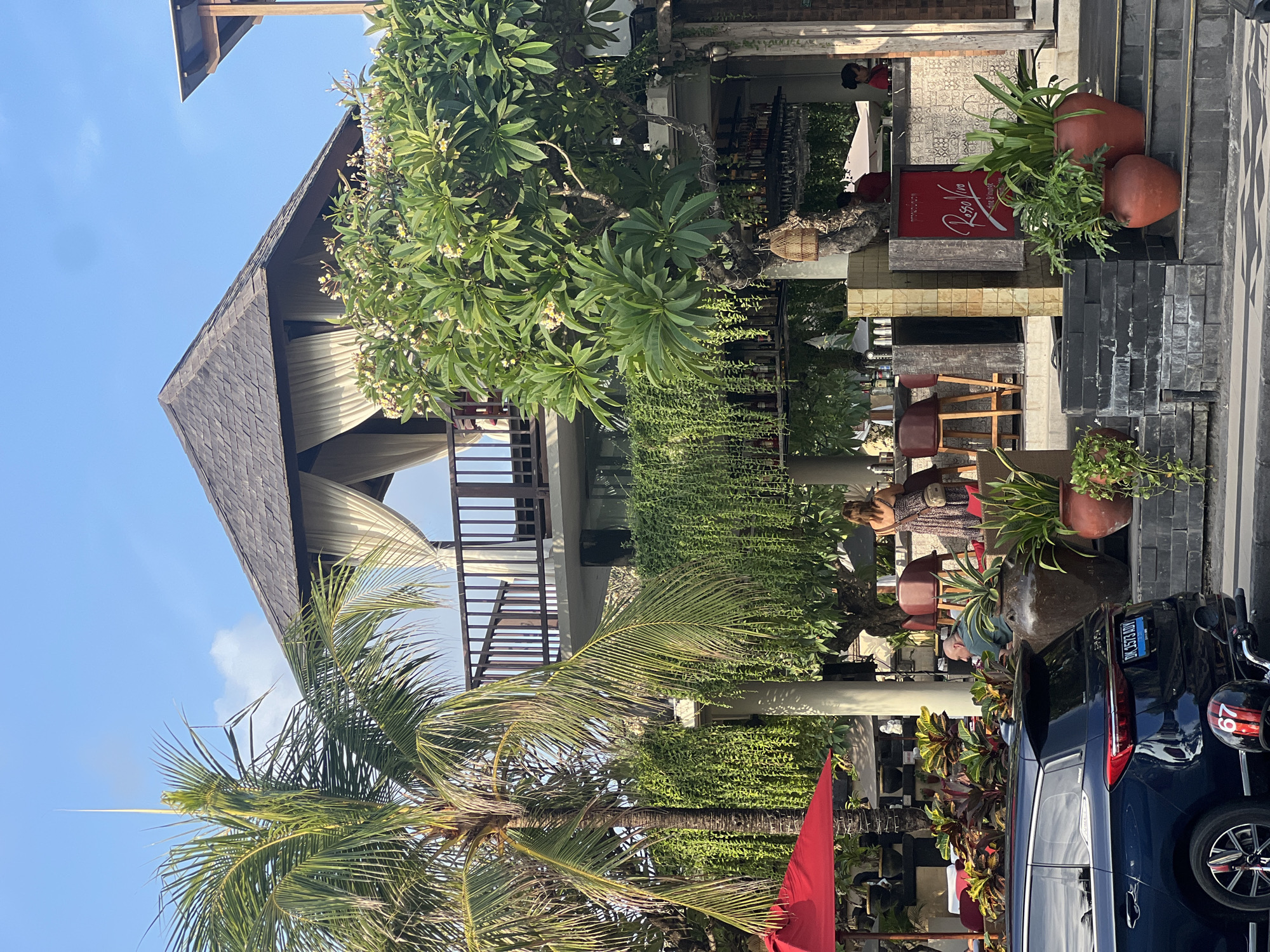 In Kuta, Indonesia, a multi-level establishment features a prominent gazebo with a dark, shingled roof and cream-colored drapes on the upper level, supported by a wooden balcony. Below this, a concrete structure houses a bar area with stools and various bottles displayed on shelves. Lush green vines cascade down from the upper levels, creating a verdant wall. To the left, a tall palm tree stands beside a red umbrella shading an outdoor seating area with plants. In the foreground, a dark blue SUV is parked, with a motorcycle helmet visible on its rear. Several individuals are present: a woman in a patterned dress is seated at the bar, another person is visible behind the bar, and two individuals in red shirts are also at the bar. The environment suggests a tropical setting with clear skies and bright sunlight.