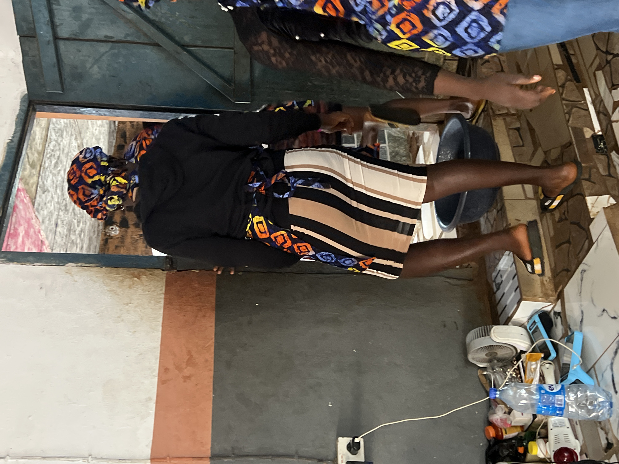 The image shows two people, likely women, in what appears to be an indoor setting, possibly a home or workshop, in Nkpor, Nigeria. The person in the foreground is wearing a black long-sleeved shirt, a headscarf with a colorful geometric pattern, and a striped skirt. They are standing next to a large dark basin, and their left leg is bent, with their foot resting on a flip-flop. The person in the background is partially visible, wearing a blue and orange patterned top and black lace sleeves, with their hands extended as if holding something or gesturing.
The scene is somewhat cluttered with various objects on the floor. To the right, there is a collection of items including a white fan, a blue device, a water bottle, and other miscellaneous objects. The walls are painted in different sections with white, orange, and grey colors. The floor is tiled with a mosaic pattern. A metal door is visible in the background.
The lighting suggests it might be daytime, but there are no strong indicators of the specific time of day or weather conditions. The overall impression is of a domestic or practical setting where some activity is taking place, though the exact nature of the activity is unclear from the image alone. The image is oriented vertically, so the description is from that perspective.