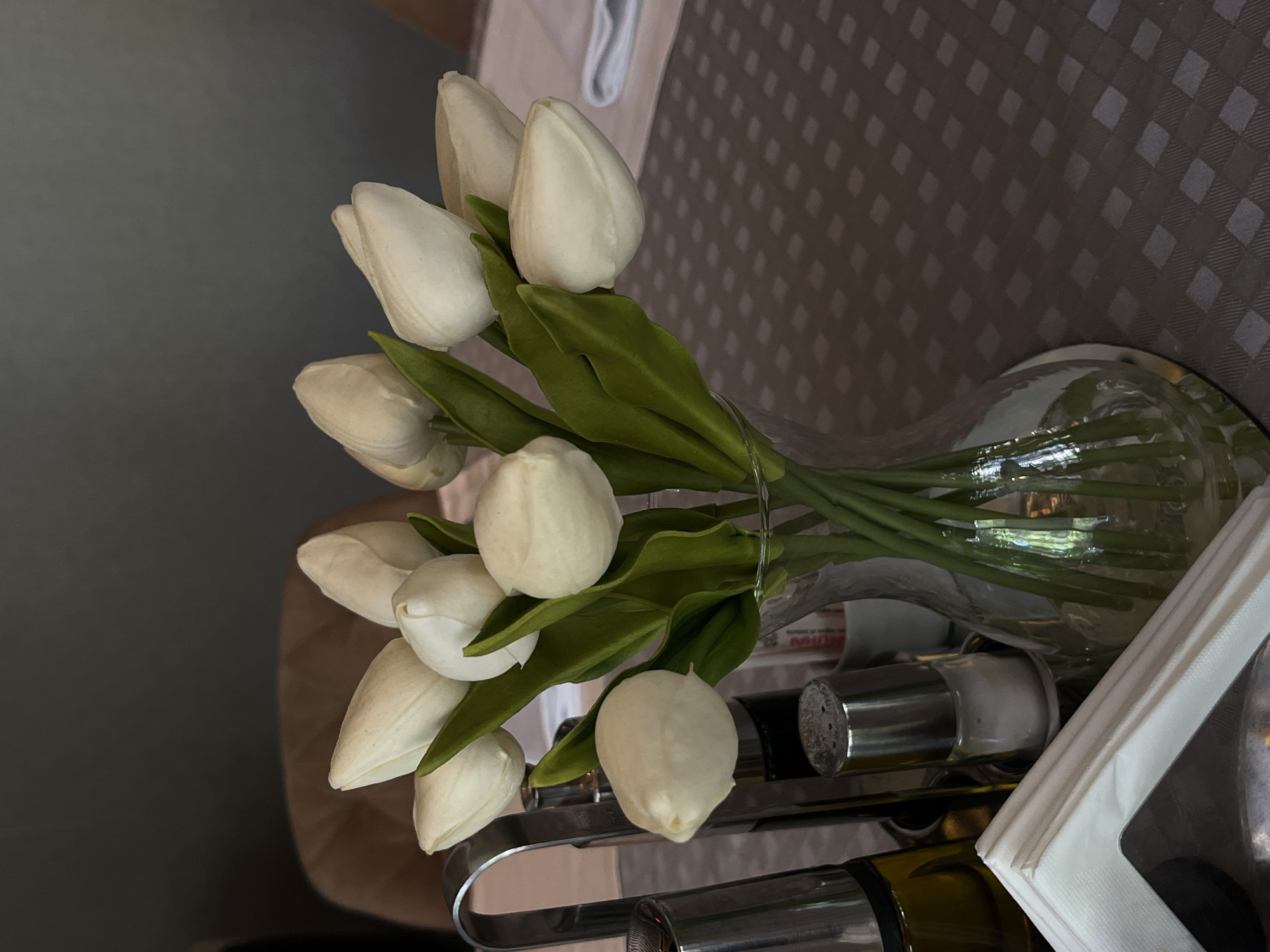 A vase of white tulips is positioned on a table. Adjacent to the vase, salt and pepper shakers, napkins, and a bottle of olive oil are present in a metal holder. The table is covered with a checkered tablecloth. The location is within Budva, Montenegro.