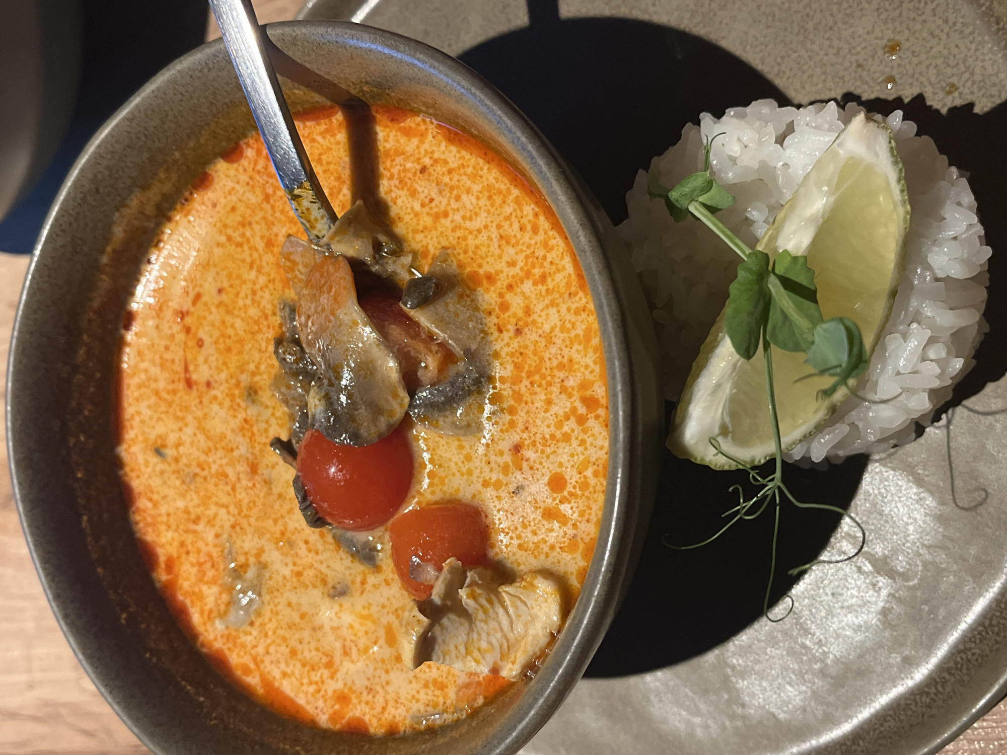 A close-up shot captures a bowl of soup with a spoon in it, next to a mound of rice with a lime wedge and sprig of greens, presented on a neutral-toned plate. The soup appears creamy, with a reddish-orange hue and visible ingredients like tomatoes, mushrooms, and chicken. The rice is white and fluffy, and the lime wedge has a bright green rind. The scene is lit with a combination of natural and artificial light, casting shadows that highlight the textures of the food. The location in the city of Unknown, Unknown. cannot be determined from the image.