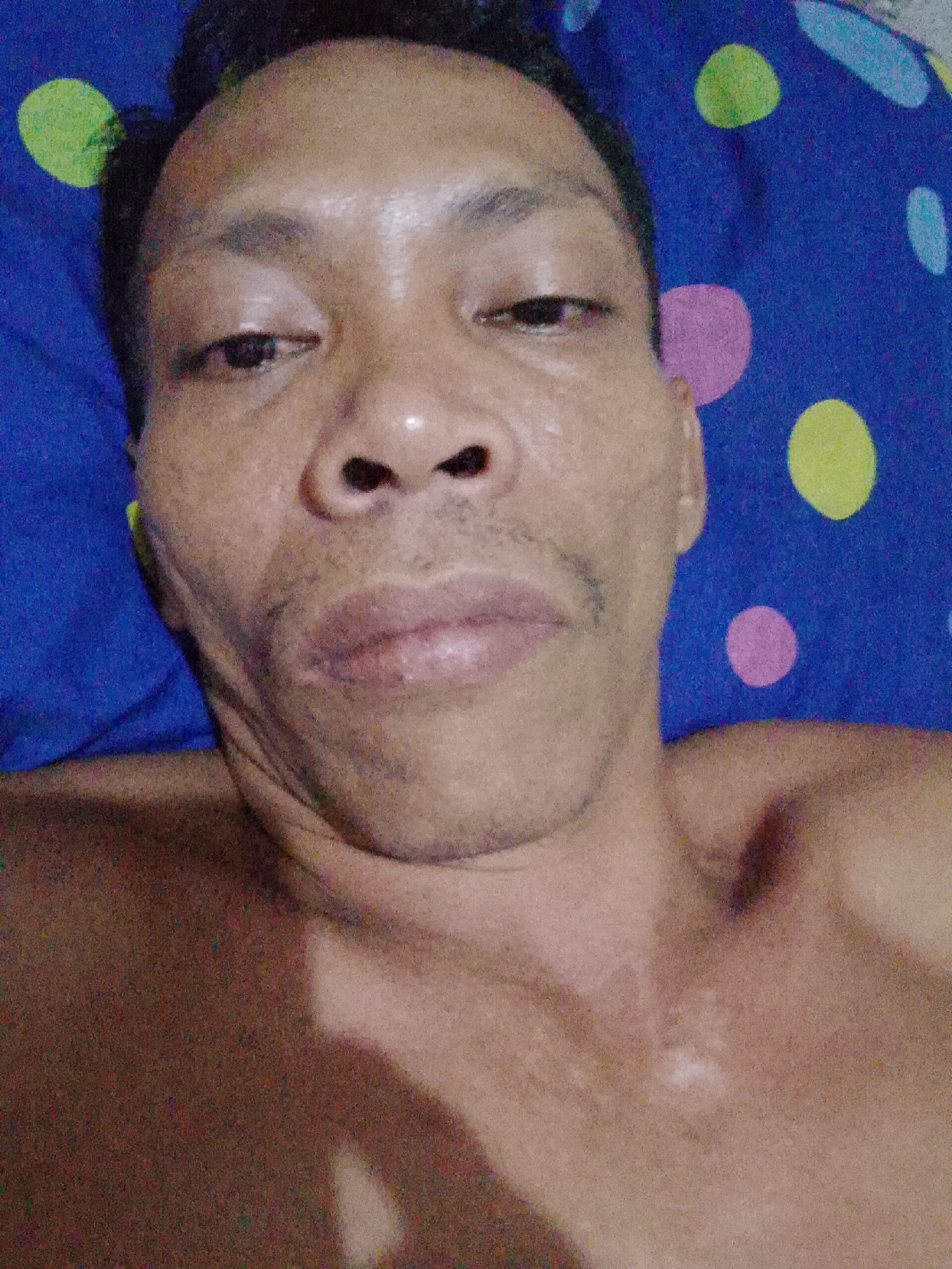 A close-up, dimly lit image shows a shirtless man lying on his back on a blue and multicolored polka-dot patterned surface. The man has short dark hair and appears to be of Southeast Asian descent. The photograph was taken from an overhead perspective. There is a shadow cast on the lower right area, and his chest area is partially visible. Based on the prompt, the image was taken in Bukit Ketok, Indonesia.