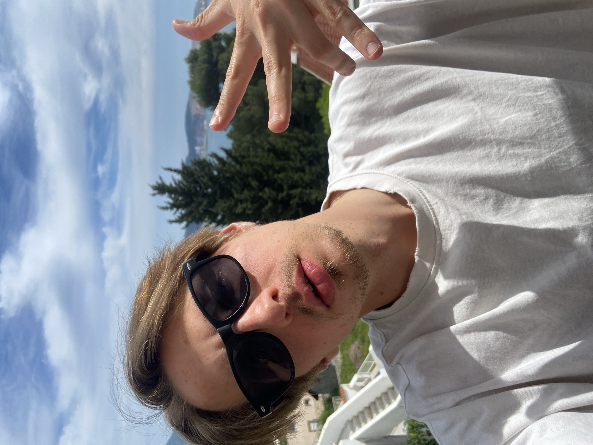 A selfie of a young, fair-skinned male with blonde hair and a sparse beard, wearing black sunglasses and a white t-shirt. He has his right hand raised, fingers spread, partially obscuring the background. Behind him is a view of the sky with scattered clouds, a body of water, and tree-covered land. A white railing is visible in the lower right corner. The location is Šušanj, Montenegro, implied by the typical vegetation and skyline of the area.