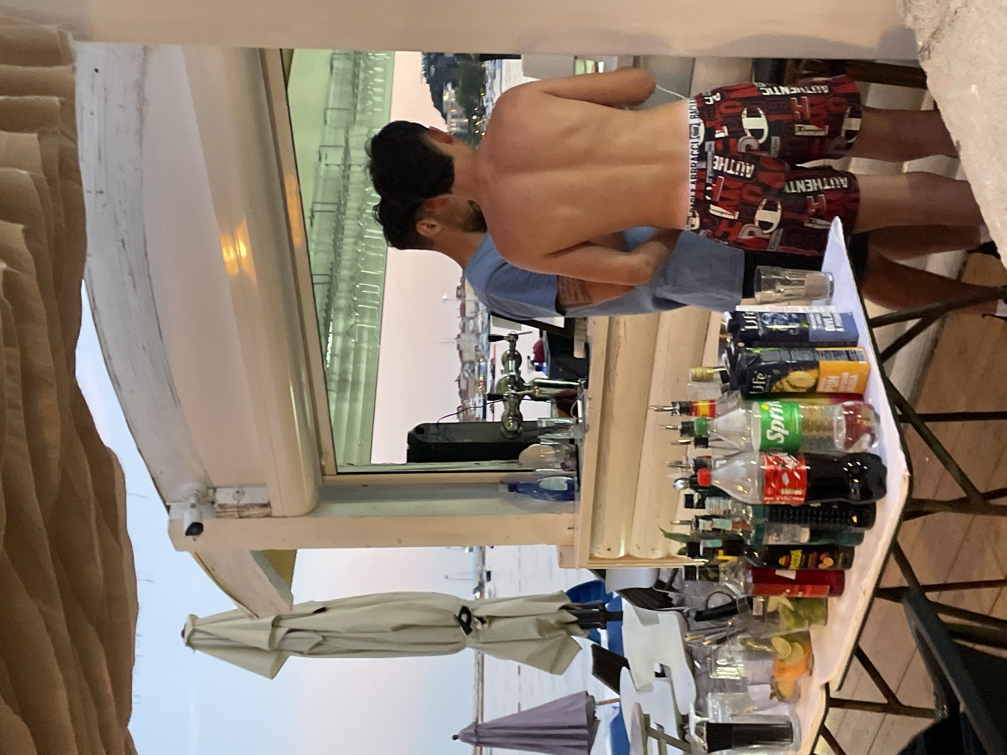 At sunset, two men are standing behind a bar setup at an outdoor establishment, possibly a beach bar, located in Budva, Montenegro. The man on the right, facing away from the camera, is shirtless and wearing patterned shorts. The man on the left is wearing a blue t-shirt. They appear to be interacting with the bar's offerings, which include a variety of bottled beverages, glasses, and bar equipment such as a beer tap. In the background, there is a body of water with boats, and a cityscape with buildings visible on a hillside. The atmosphere suggests a relaxed, evening setting.
