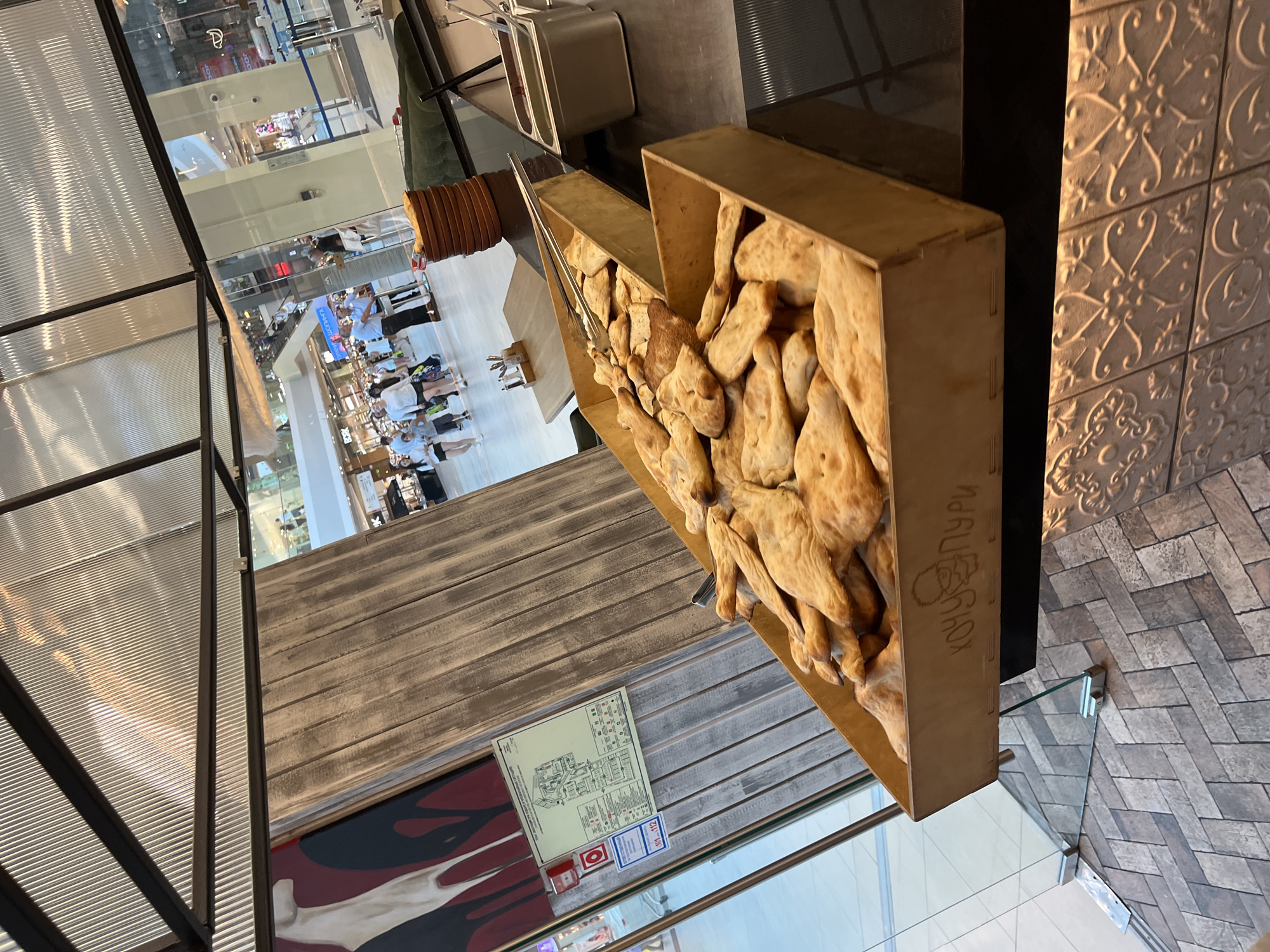 In a brightly lit indoor setting, possibly a food court in Sochi, Russia, a wooden bin overflowing with freshly baked flatbread is prominently displayed. The bread, golden brown and puffy, is stacked and piled high, with a pair of metal tongs resting on top, suggesting self-service. To the right, a metal container with a lid holds a green sauce, likely a condiment. In the background, through large glass windows, shoppers can be seen strolling through a mall, some near escalators. A wooden paneling with a vertical grain texture lines the wall behind the bread display. On this wall, a fire safety plan is mounted, along with a red fire alarm. The floor beneath the bread bin is tiled in a herringbone pattern, and further in the foreground, a decorative tiled surface with an ornate relief pattern is visible. The overhead lighting is diffused through a corrugated metal ceiling. The overall scene depicts a food vendor in a public space, likely a mall, in Sochi.
