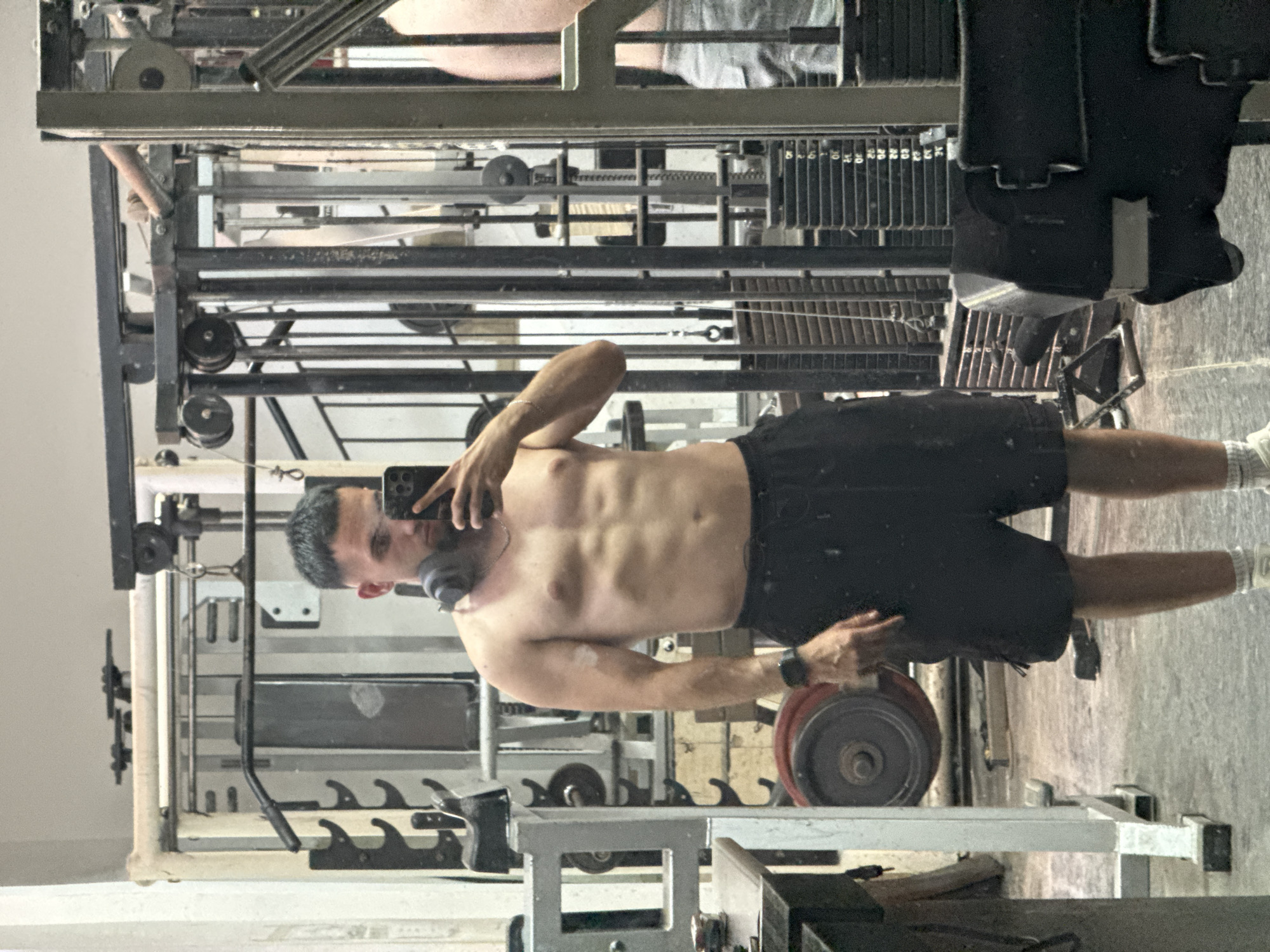A man with short, dark hair and headphones around his neck stands in a gym, taking a mirror selfie with his phone. He is shirtless, wearing black shorts and white socks. His abdominal muscles are visible. The gym appears to be a weightlifting facility with various machines, weight racks, and weight plates. In the background, another person wearing grey shorts is reflected in a mirror, partially visible. The overall environment suggests a functional, perhaps older, gym setting.