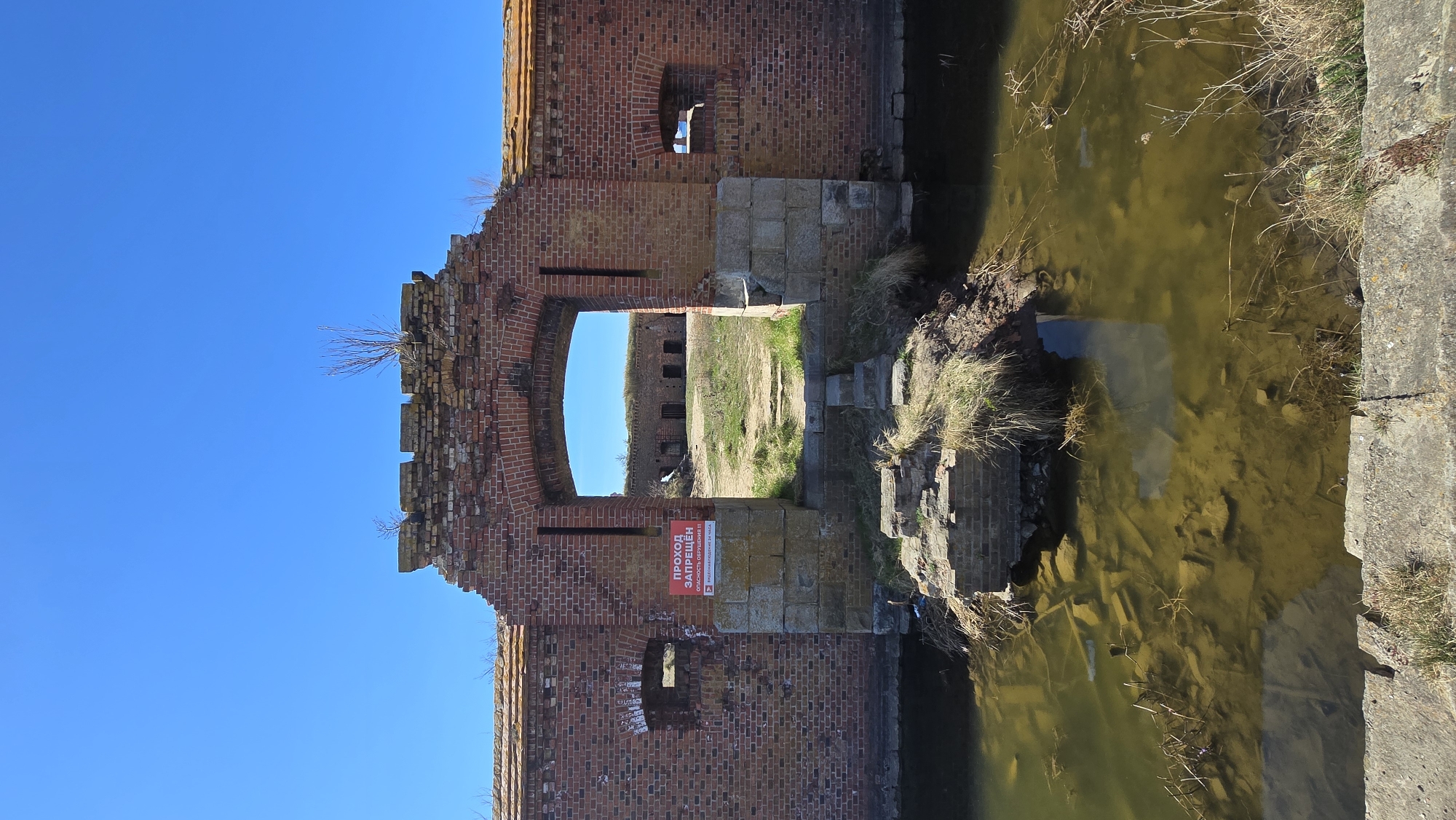 The image shows a ruined brick structure, possibly a fort or building remnant, in Baltiysk, Russia. The ruins frame a view of a grassy area and another ruined structure in the distance. A body of water, seemingly a moat or canal, is present on one side of the structure, with submerged debris visible within it. A sign in Russian hangs on the brick structure. The sky is clear and blue.