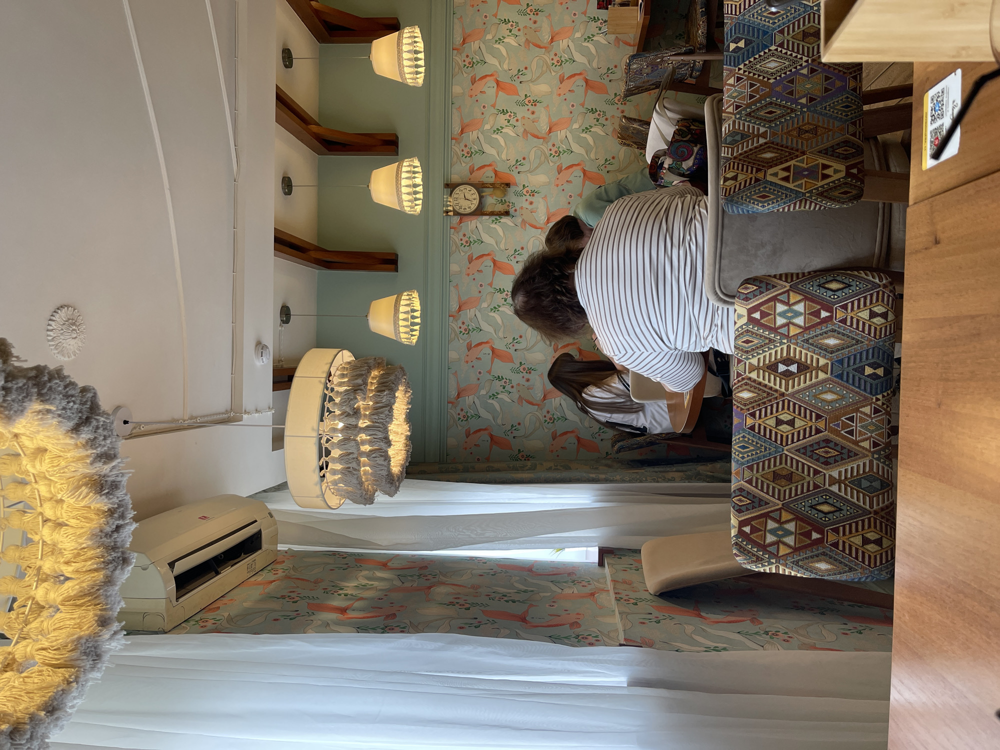 The image depicts an interior scene, likely a cafe or restaurant, with several patrons seated at tables. The decor features a distinctive wallpaper with a repeating pattern of orange and white stylized figures against a light blue background. Three pendant lamps with fringed trim hang from the ceiling, casting a warm light. An air conditioning unit is visible on the wall above sheer white curtains. In the foreground, a wooden table is partially visible, along with chairs upholstered in a colorful, geometric fabric. A clock hangs on the wall in the background. The scene suggests a casual dining or social gathering. The location is identified as Izhevsk, Russia.