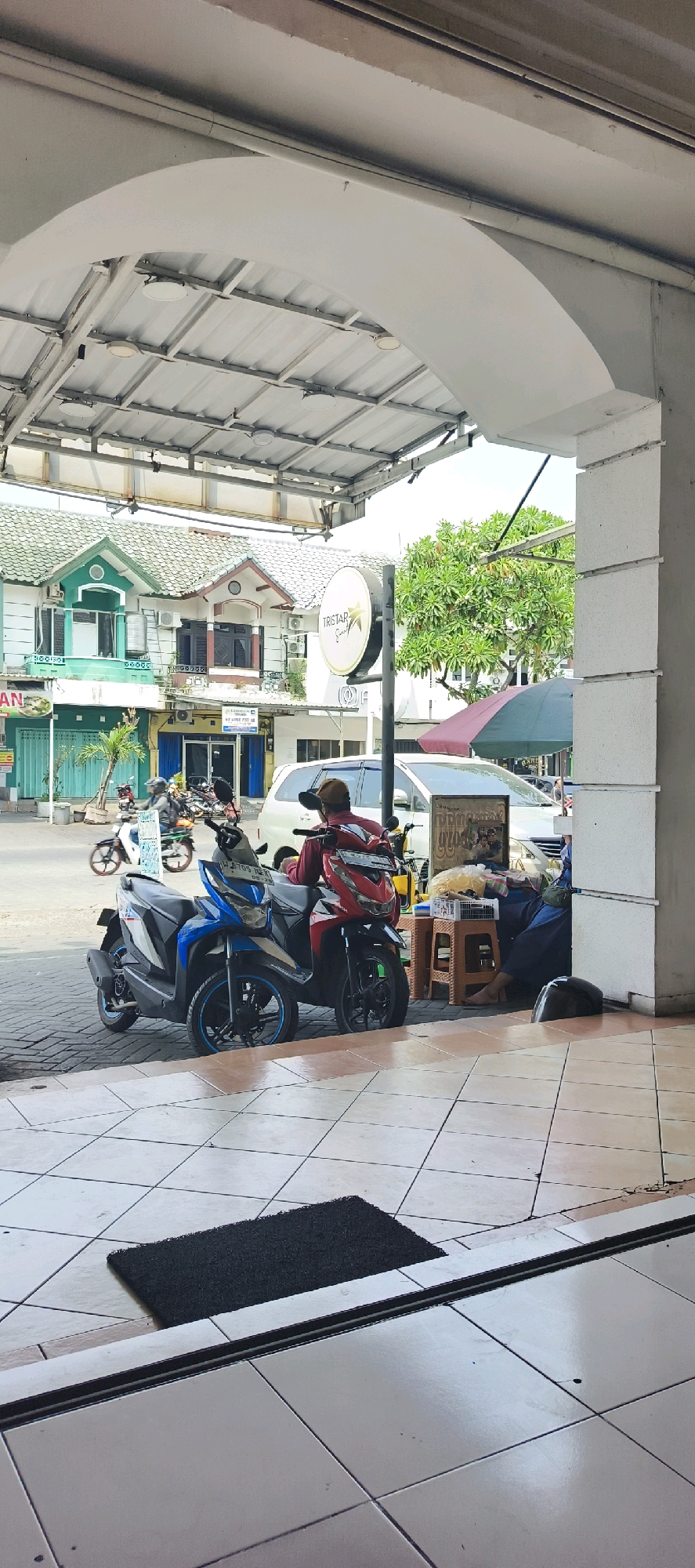 In the city of Kureksari, Indonesia, a street-level view captures a modest commercial area under an arched awning. Two motorcycles, one blue and one red, are parked on a paved sidewalk. A person wearing a red shirt and a brown cap sits on the red motorcycle, facing away from the camera. To the right of the motorcycles, a woman in a dark blue outfit sits on a low plastic stool next to a small table displaying various items, including a woven mat and what appears to be food. Behind her, a white van is parked. The background features a row of buildings with green and white facades, some with awnings and air conditioning units. A "TRISTAR" sign is visible on a pole. A lush green tree stands to the right of the buildings. The foreground shows a tiled floor with a black doormat. The scene depicts a typical urban street scene with people engaged in daily activities.